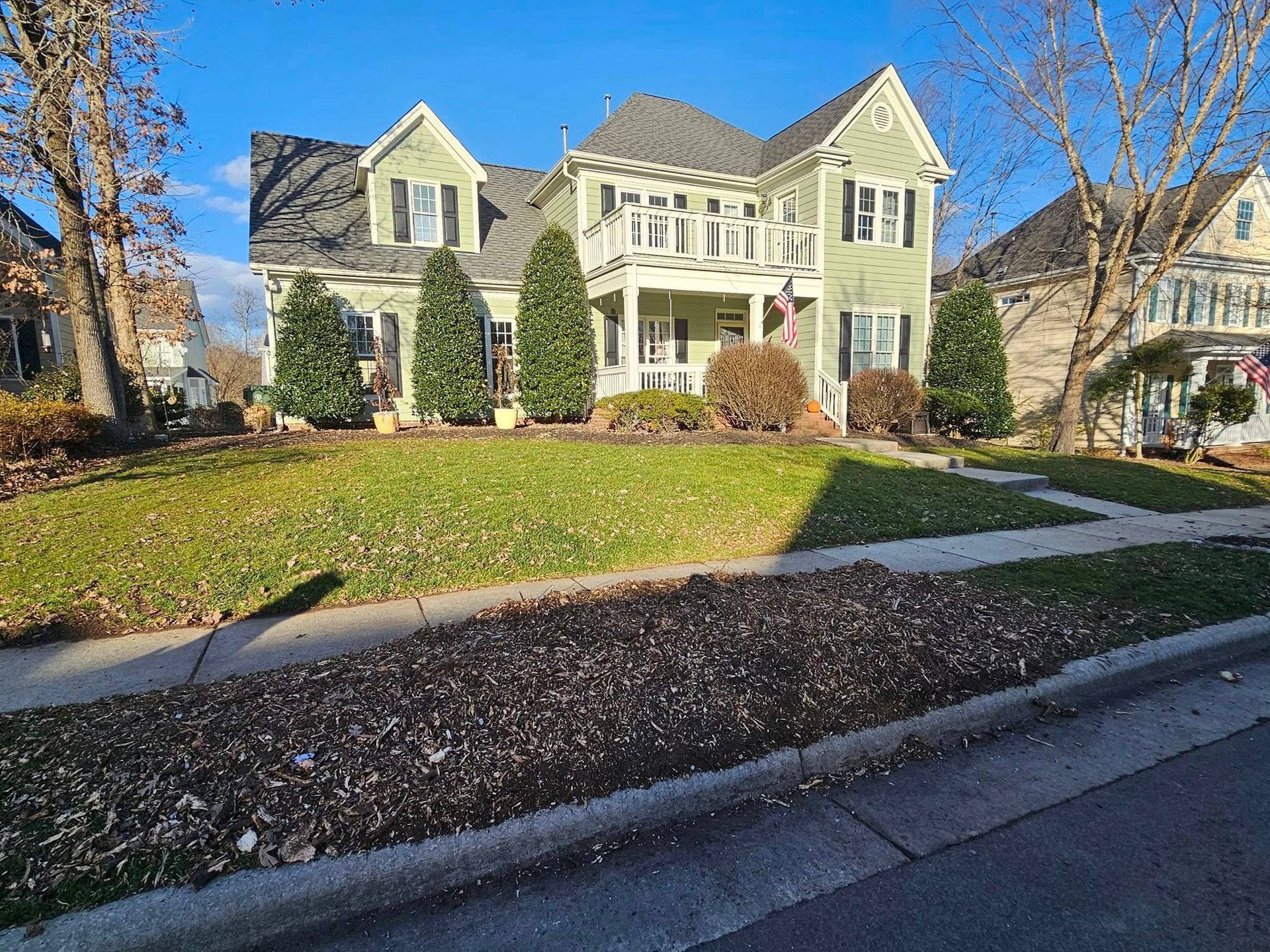 Two-story green house with a porch and bushes on a sunny day.