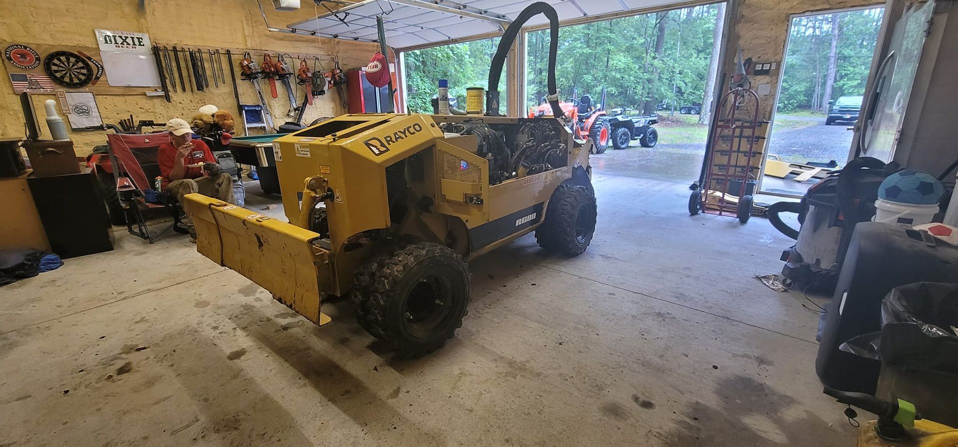 A yellow stump grinder sits inside a garage with open doors, tools on the walls, and a person in the background.