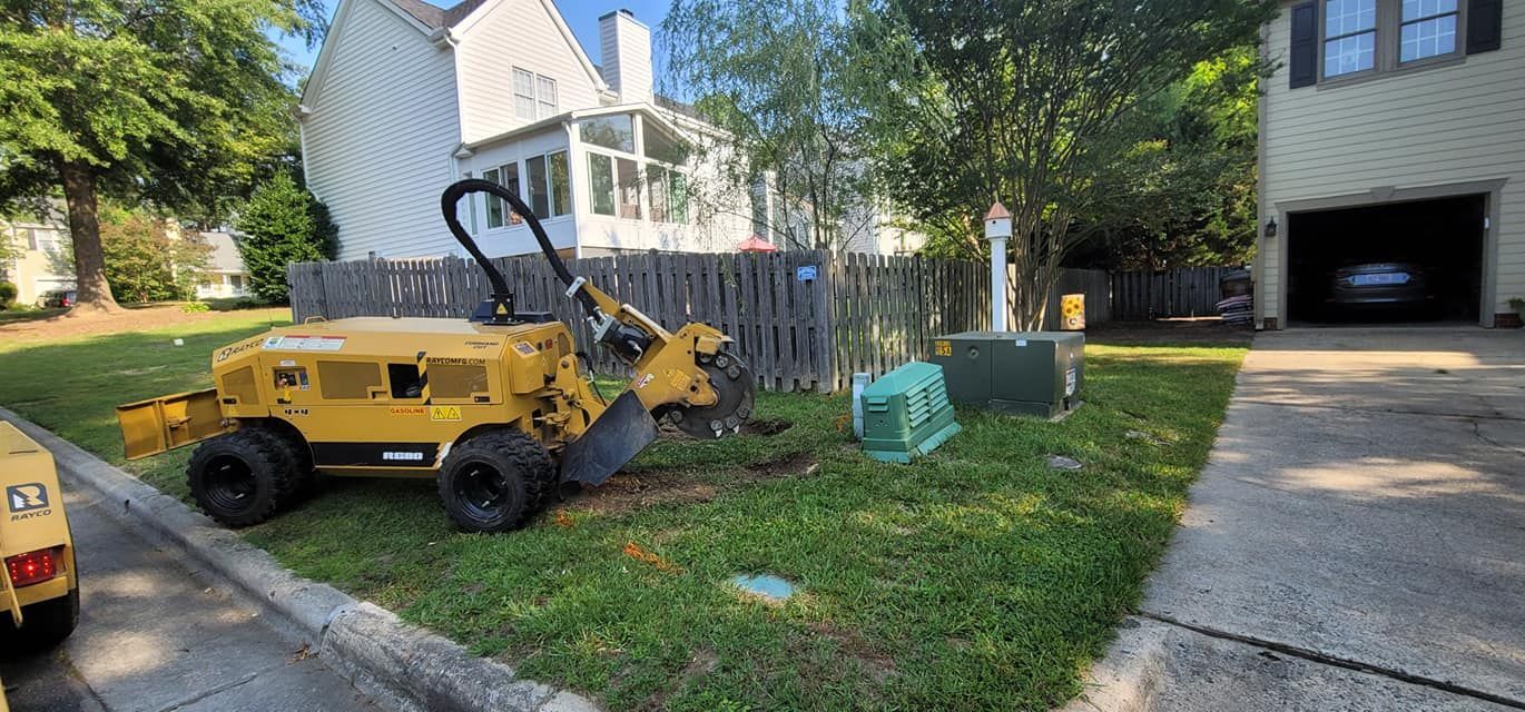 Yellow stump grinder on a residential lawn, grinding near a wooden fence and utility boxes.