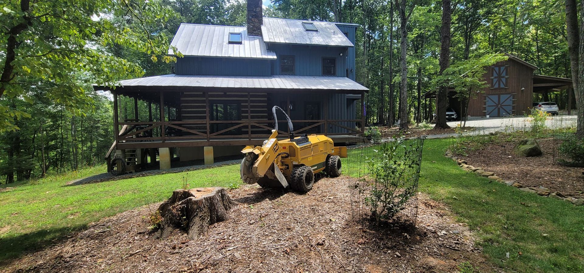A yellow stump grinder working on a hillside in front of a blue house with a porch and trees.