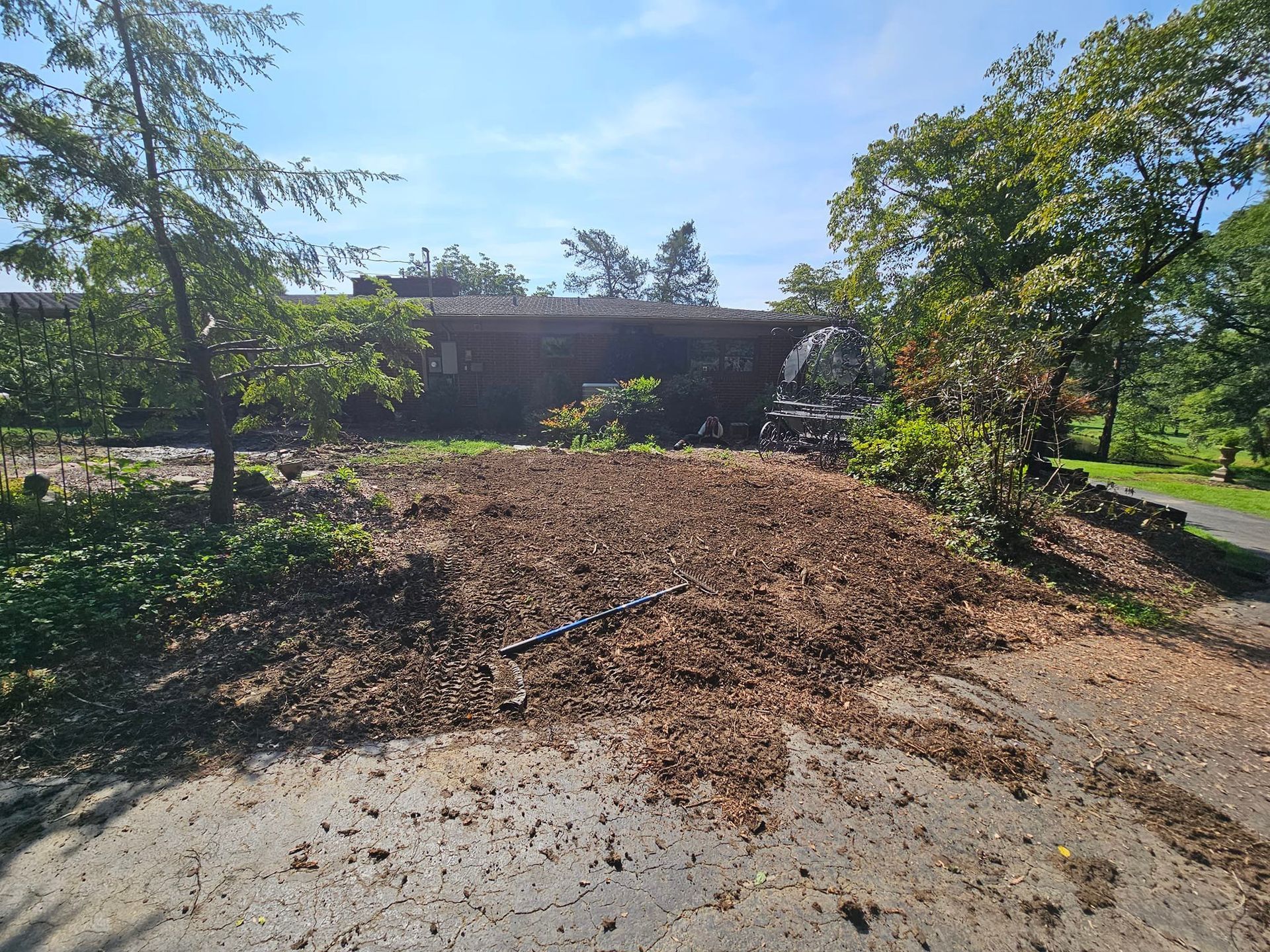Pile of mulch on a concrete surface, with trees and a brick building in the background. Bright sunny day.