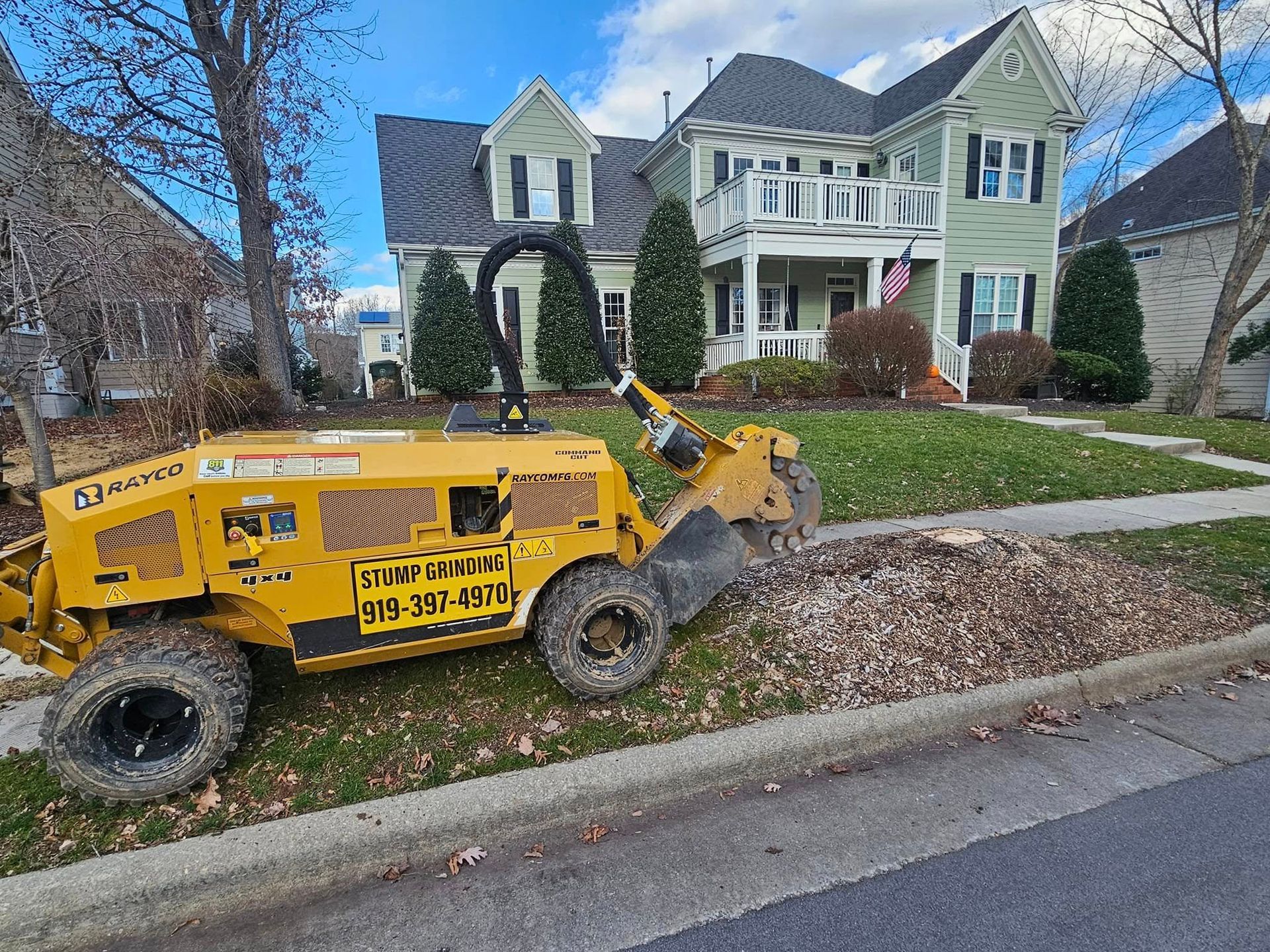 Yellow stump grinder on a residential lawn, grinding wood debris near a sidewalk and house.