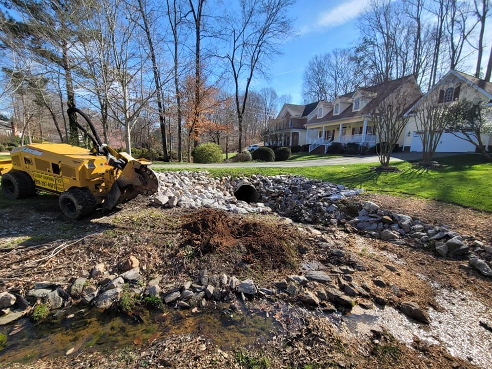 Yellow construction vehicle near a rocky drainage area with a culvert. Houses and trees in the background.