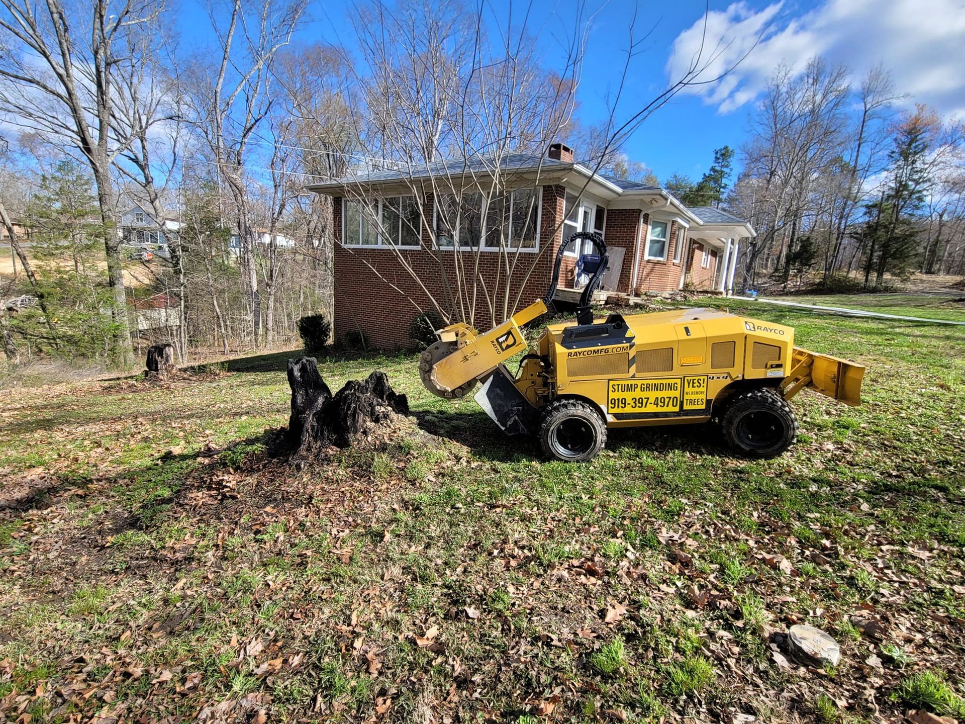 A yellow stump grinder working on a tree stump in front of a brick house on a sunny day.
