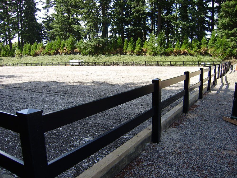 A black vinyl fence surrounds a dirt field with trees in the background