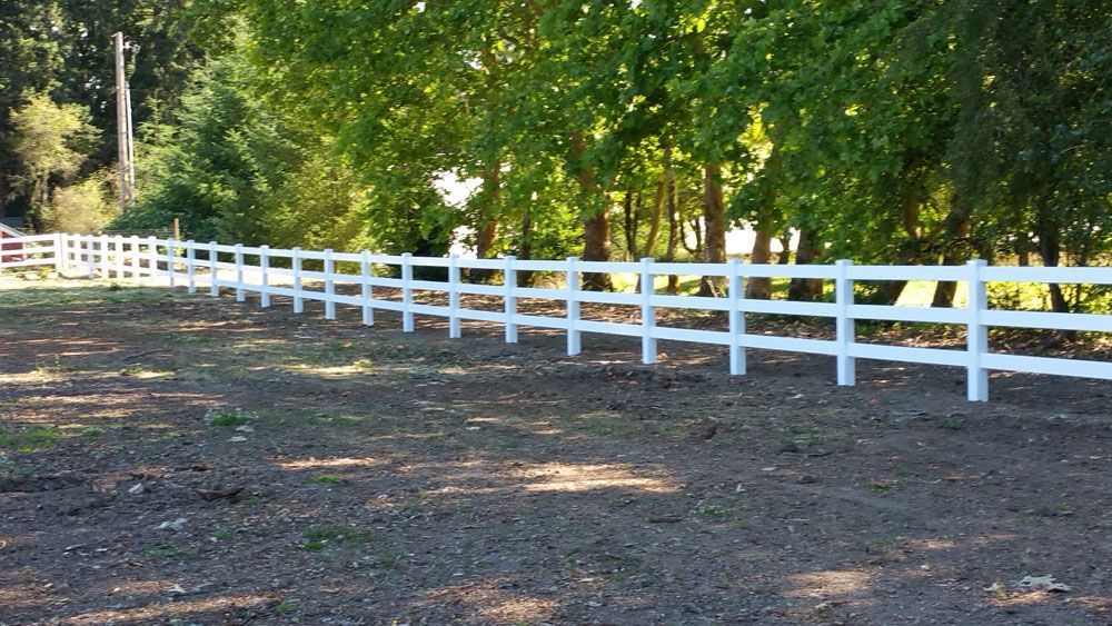 A white fence surrounds a dirt field with trees in the background