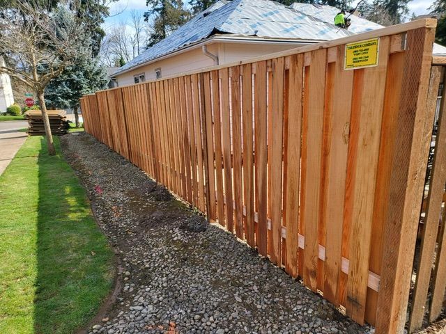 A wooden fence is sitting next to a gravel driveway in front of a house