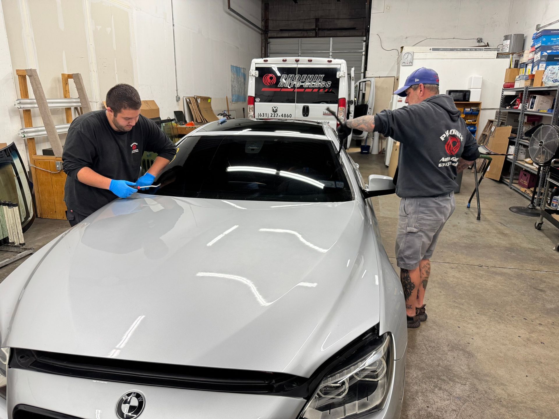 Two men tinting a silver BMW's windshield in a shop. One applies film, the other holds a tool.