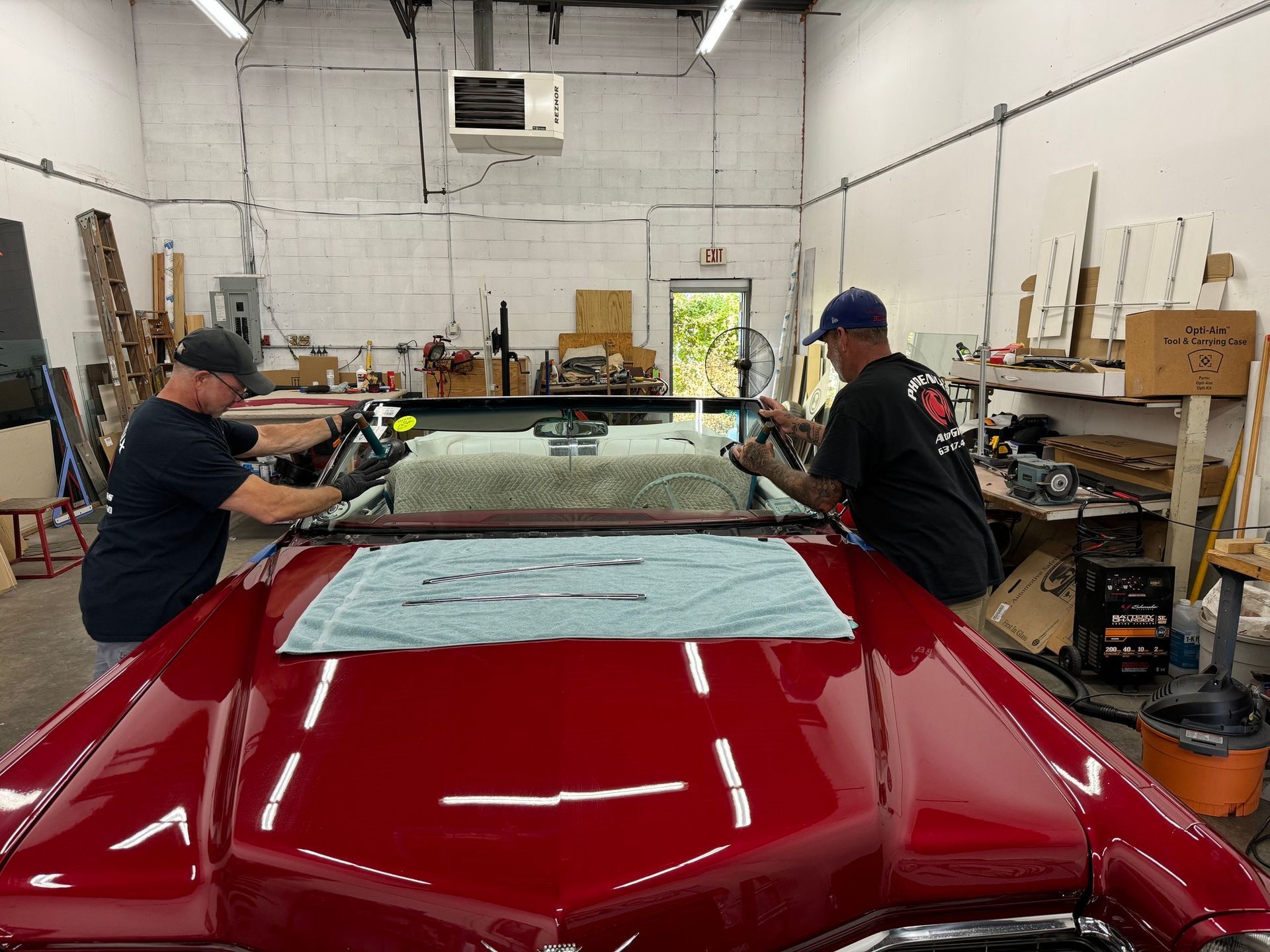 Two men installing windshield on a red classic car in a workshop.
