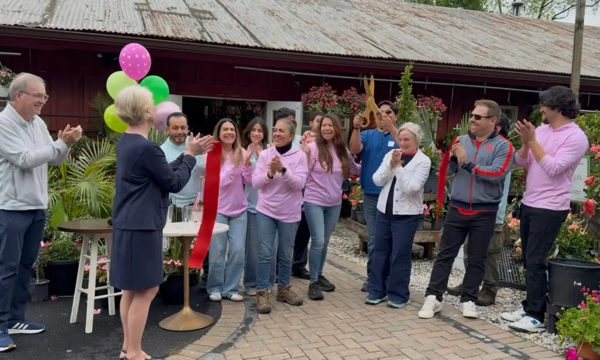 Group applauding at an outdoor celebration, with a man holding pink and green balloons near a pavilion.