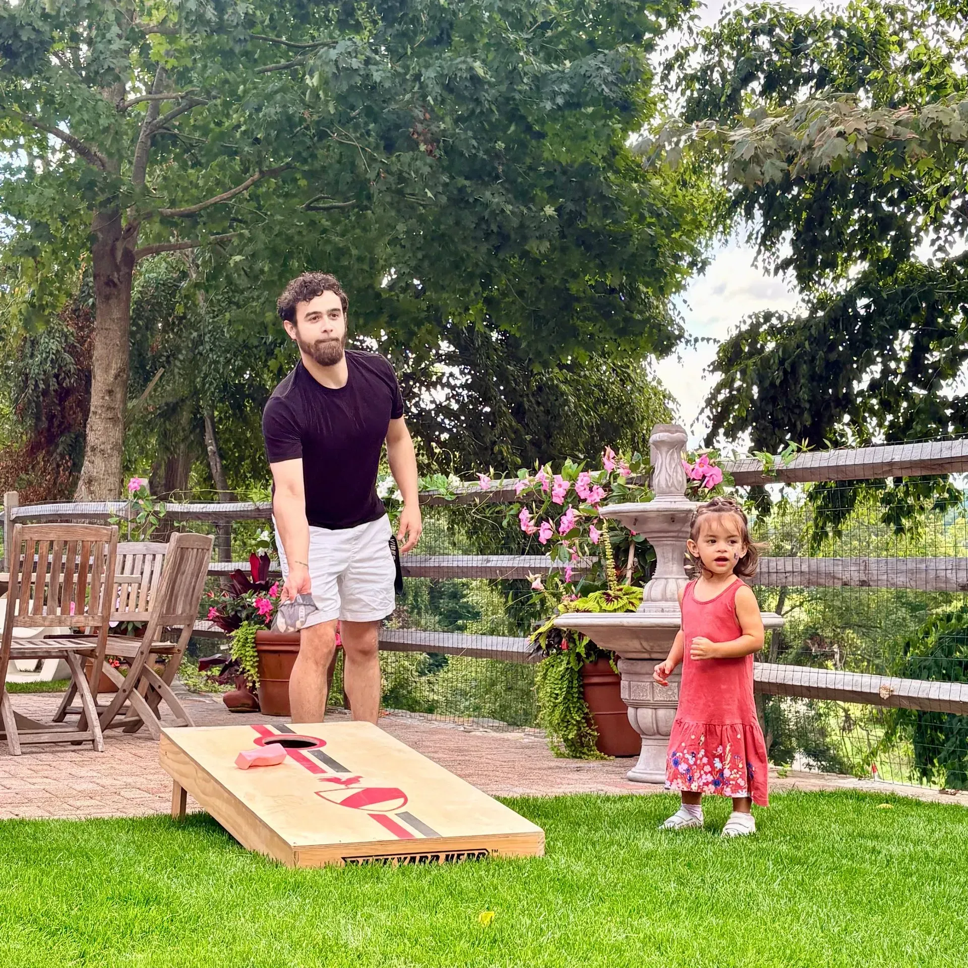 Man and child outdoors near a wooden ramp on a lawn, with trees and flowers in the background.