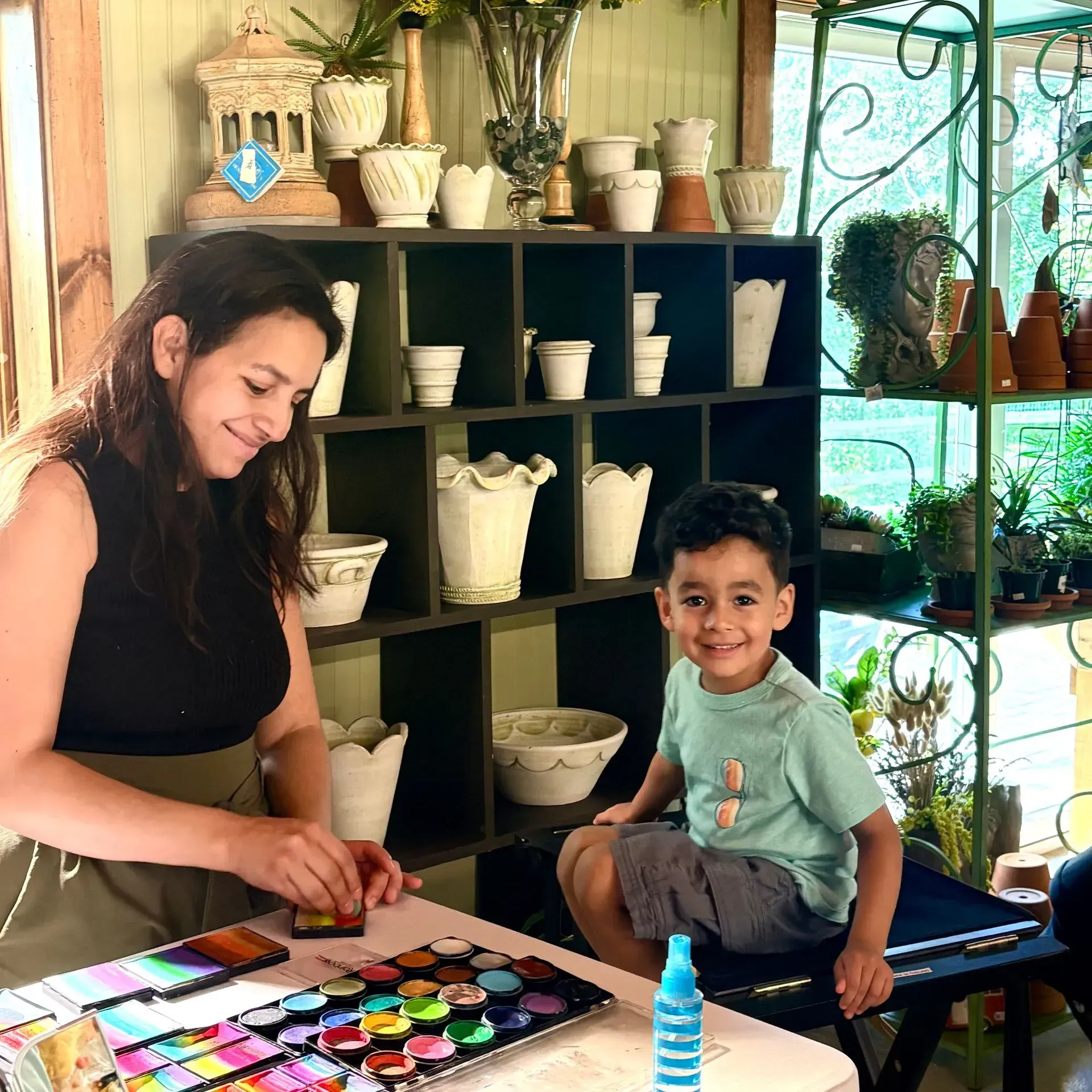 Woman and child painting at a table in a cozy art studio, with shelves of ceramic pots behind them.