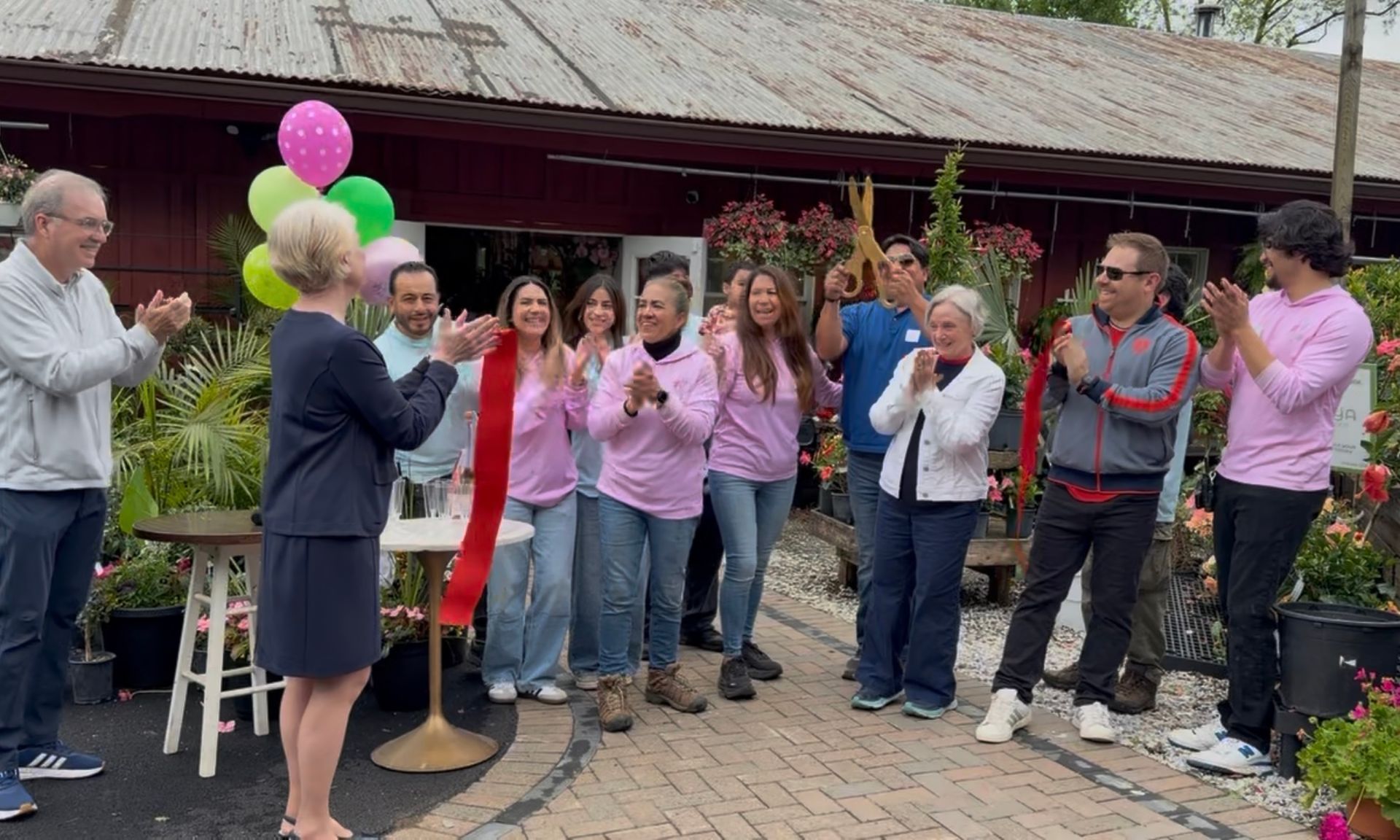 People in pink shirts and a man with balloons clap beside a doorway at an outdoor gathering.
