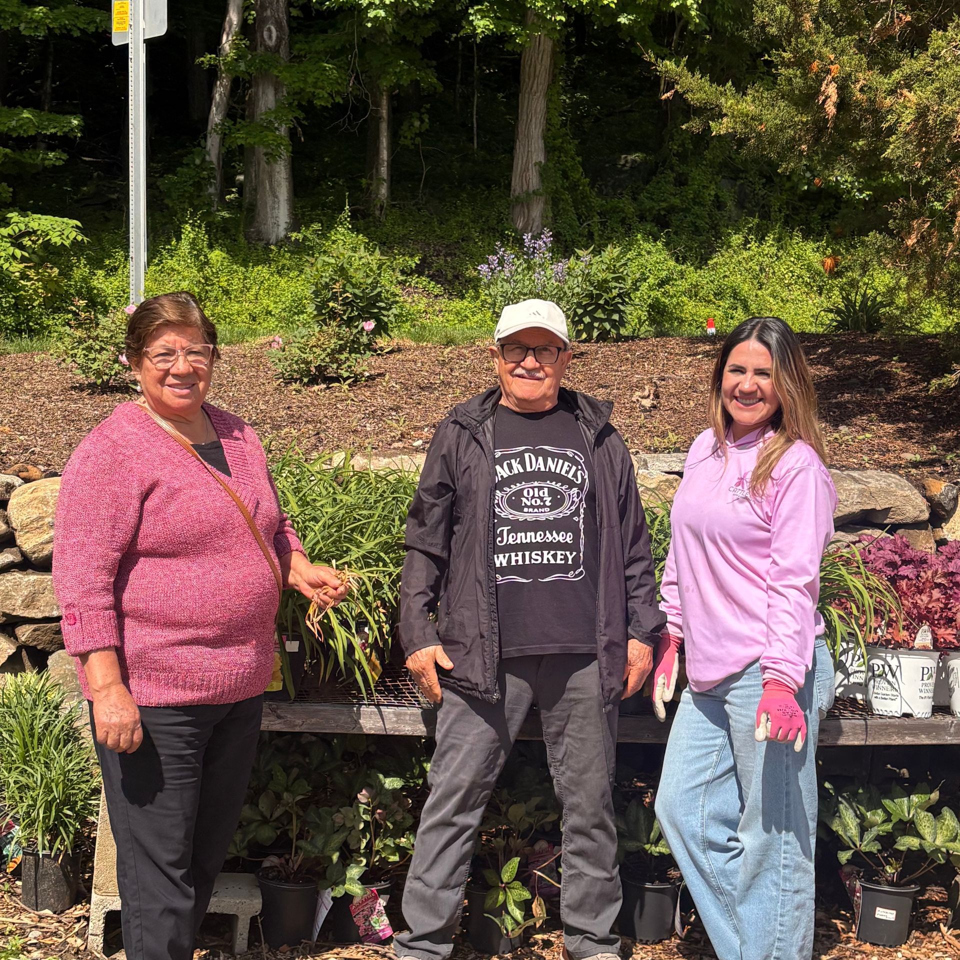 Three people posing outdoors in a sunny garden center among potted plants.