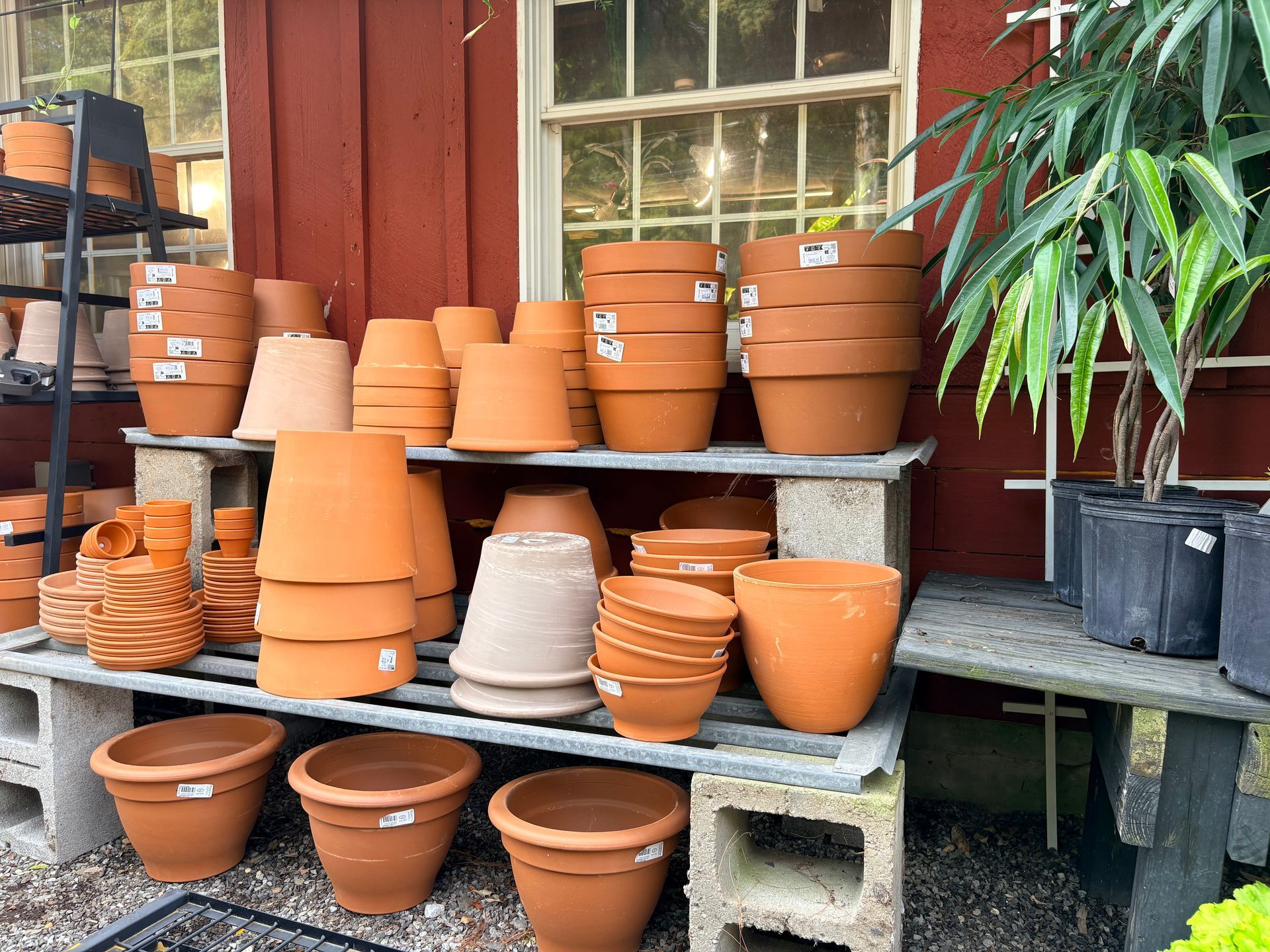 Stacks of terracotta pots and planters outside a red storefront beside a window and potted plant.