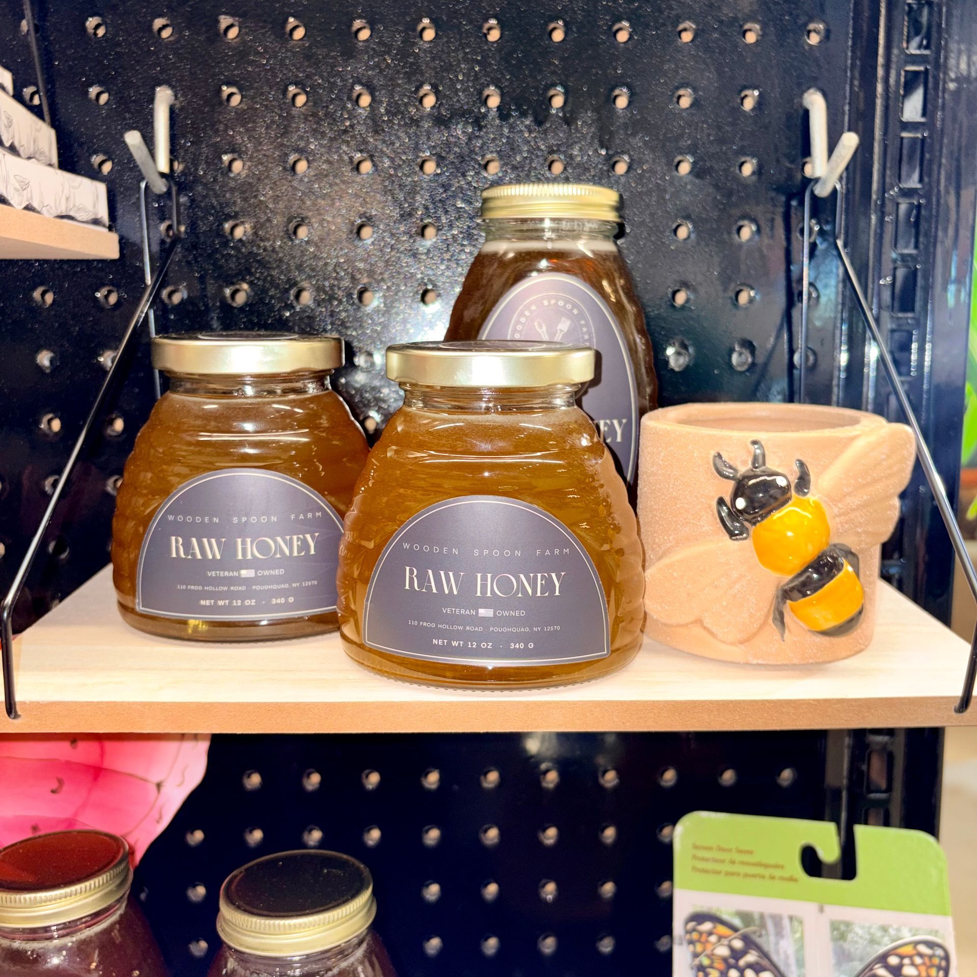 Glass honey jars and a bee-themed mug on a store shelf against a black pegboard backdrop