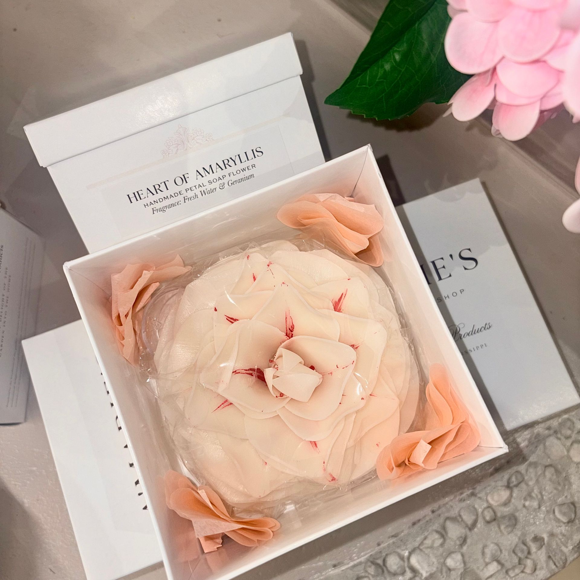 Open gift box with a cream flower-shaped soap, peach petals, and floral packaging on a gray surface.