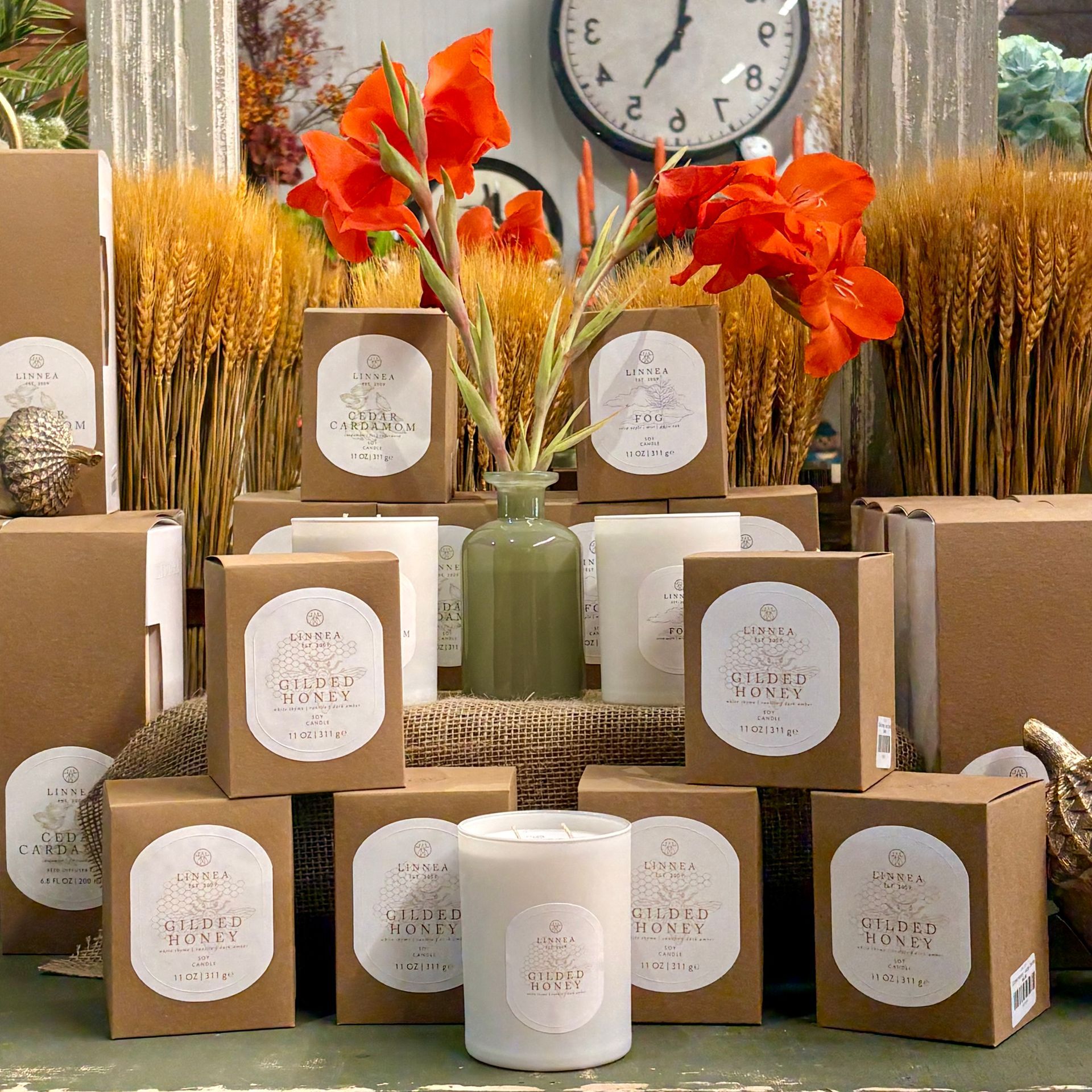 Fall display of candles and boxed gifts with orange flowers and wheat, arranged beneath a wall clock.