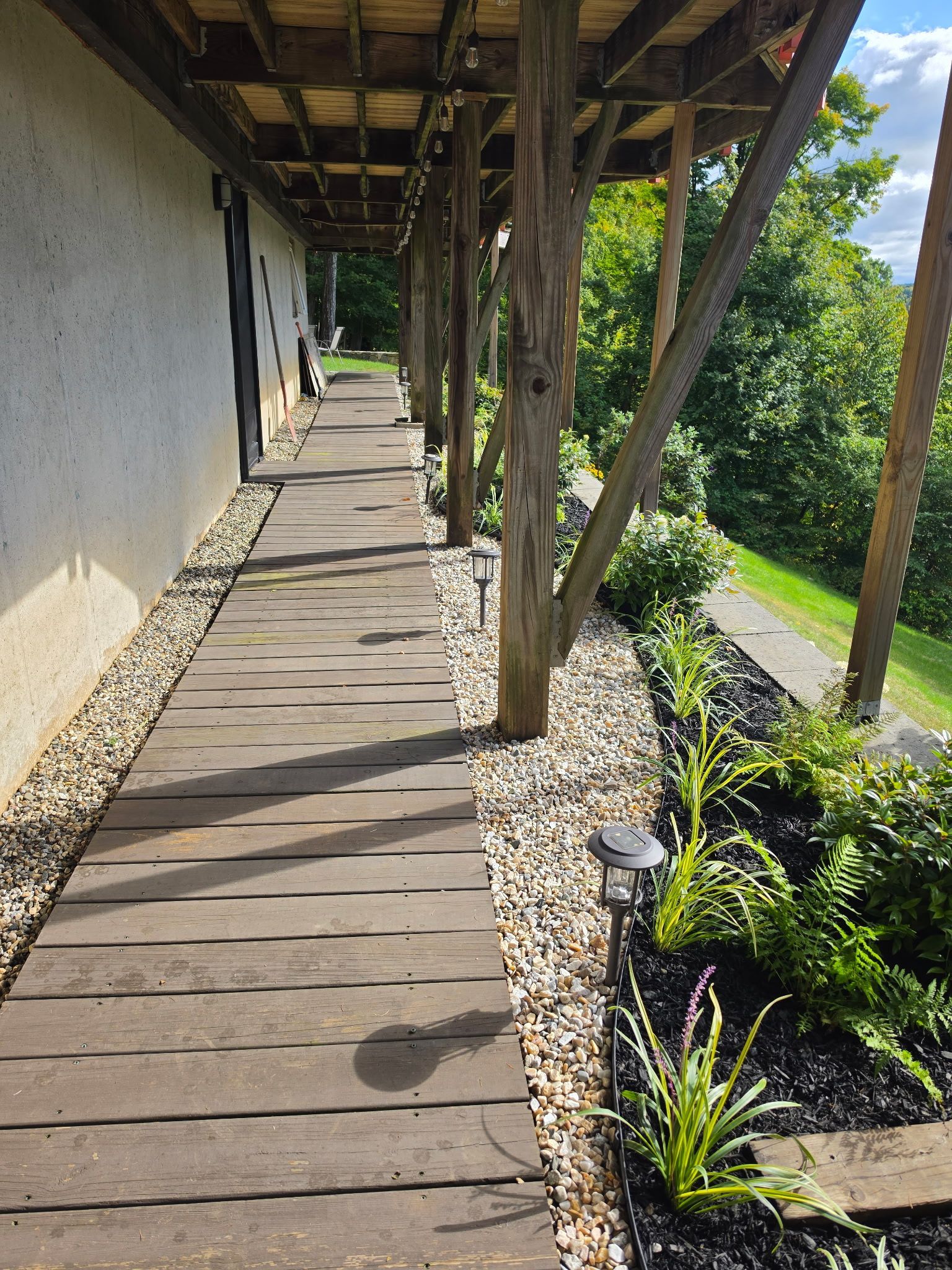 Wooden walkway beside a stucco building, with wooden posts and lush greenery below.