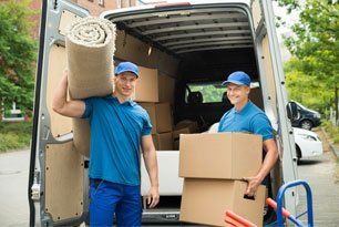 Two Male Workers Carrying Carpet And Cardboard Boxes In Front Of Van