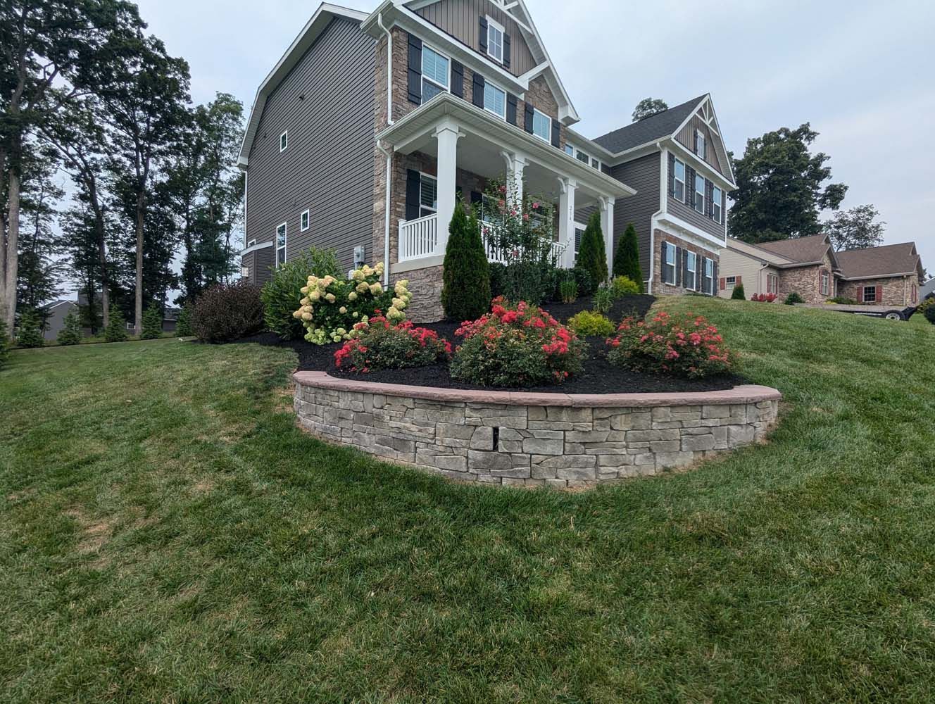 A two-story house with a brick retaining wall and a flower bed on a grassy hill.