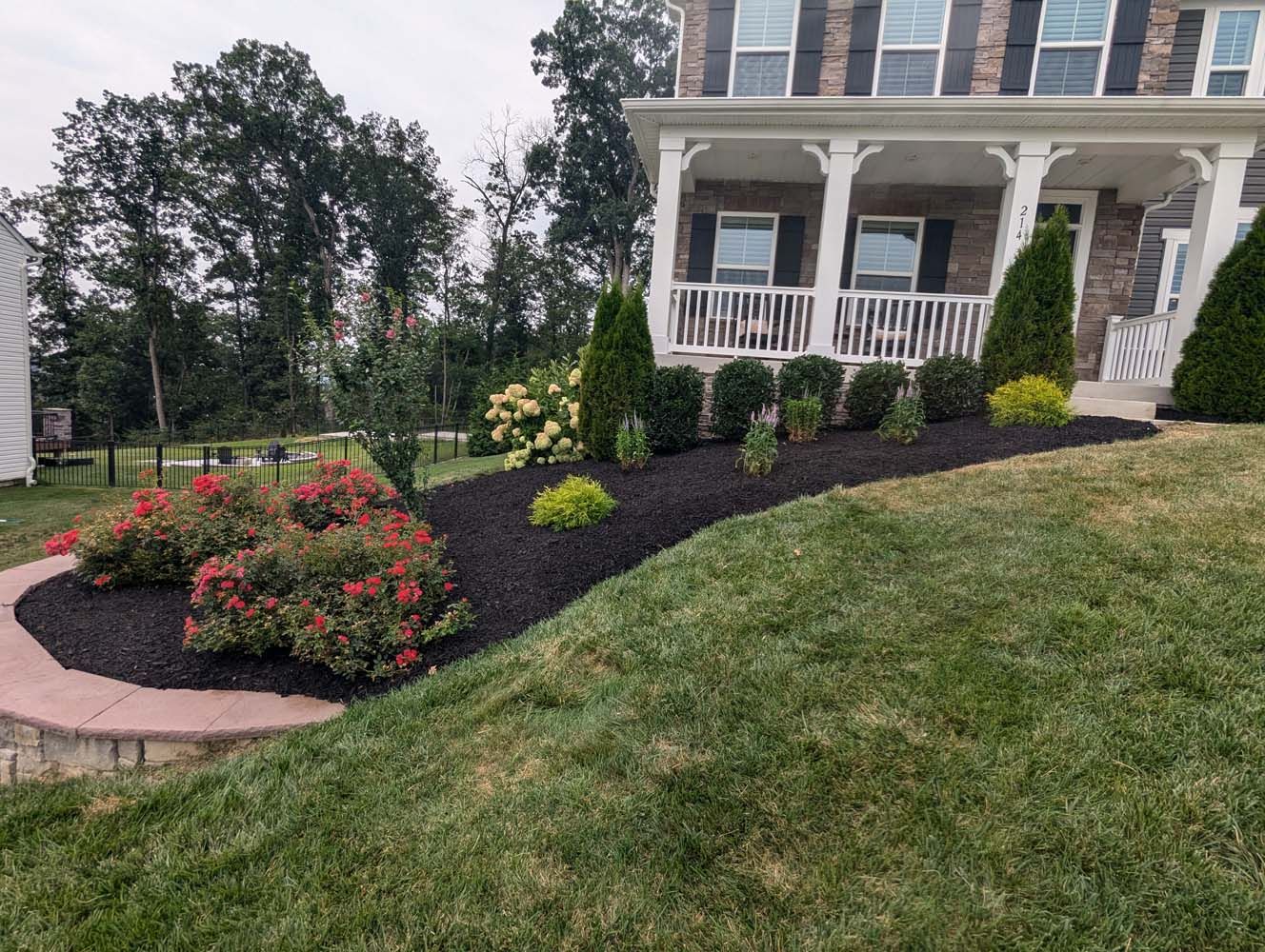 A well-manicured front yard with a brick-lined flower bed filled with bushes and dark mulch, next to a grassy lawn.