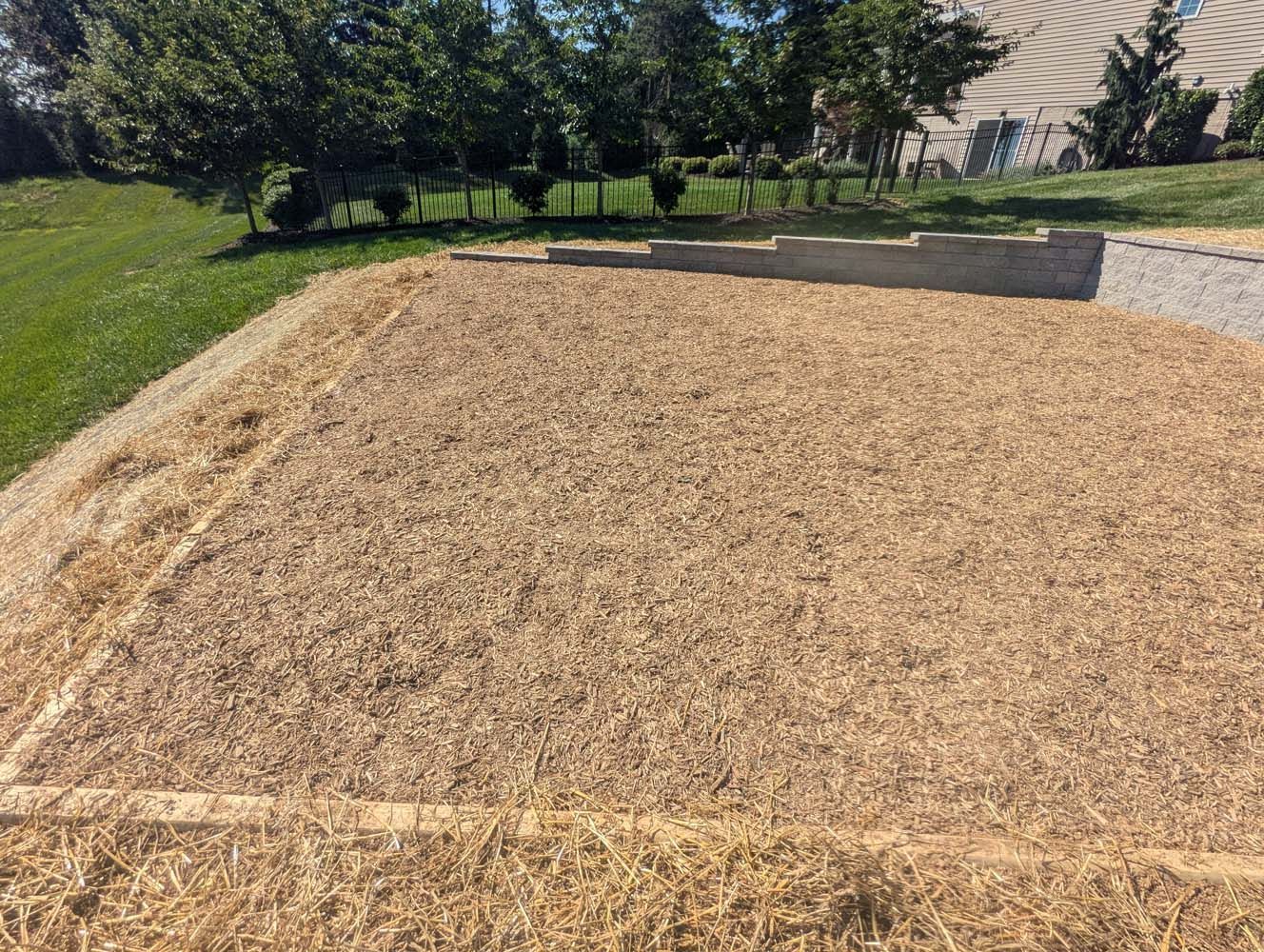 A patch of land covered in straw, next to grass and a retaining wall, under a sunny sky.