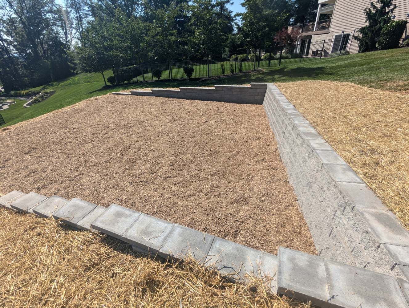 A level area with brown wood chips, contained by gray block retaining walls and steps, on a grassy hillside.