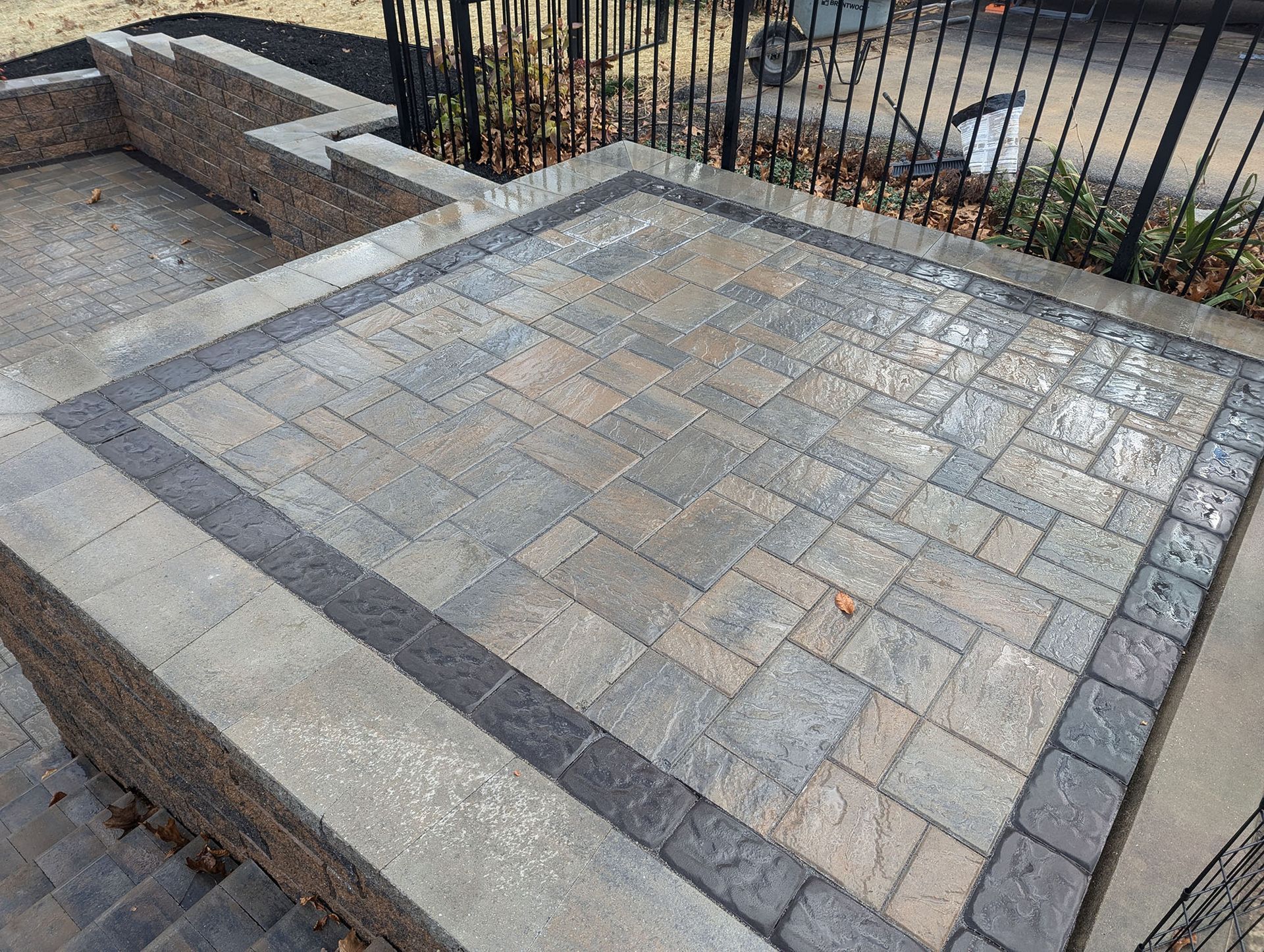 Paver patio with varying gray tones, bordered by darker pavers and retaining walls, near a black metal fence.