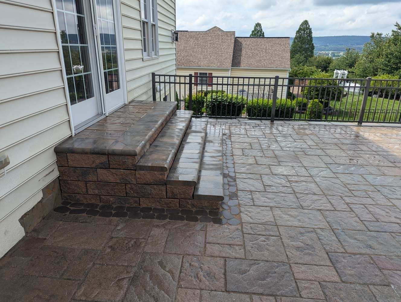Brick steps lead to a patio with a metal railing, adjacent to a light-colored house.