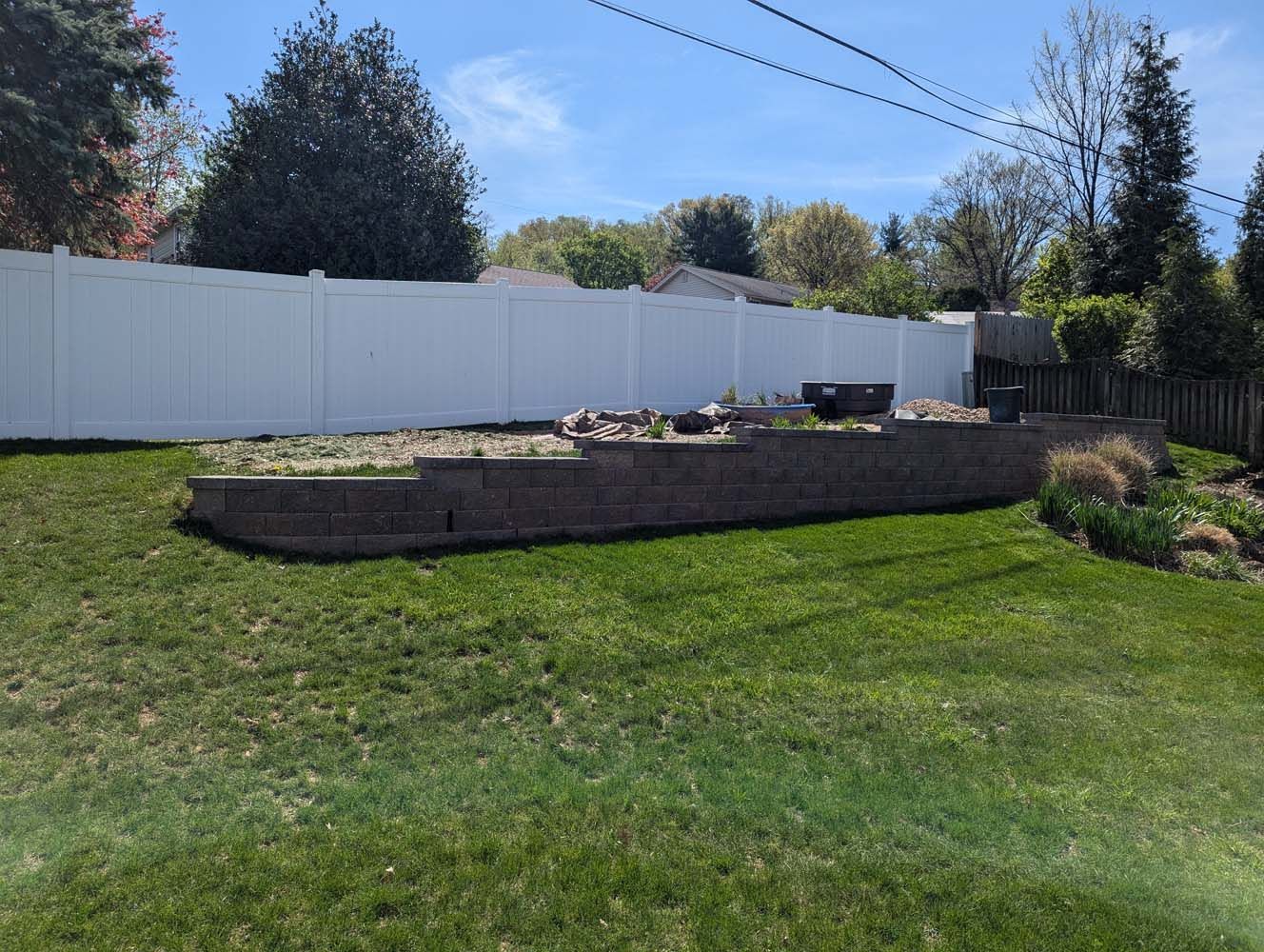 A backyard with a retaining wall, white fence, and green grass on a sunny day.