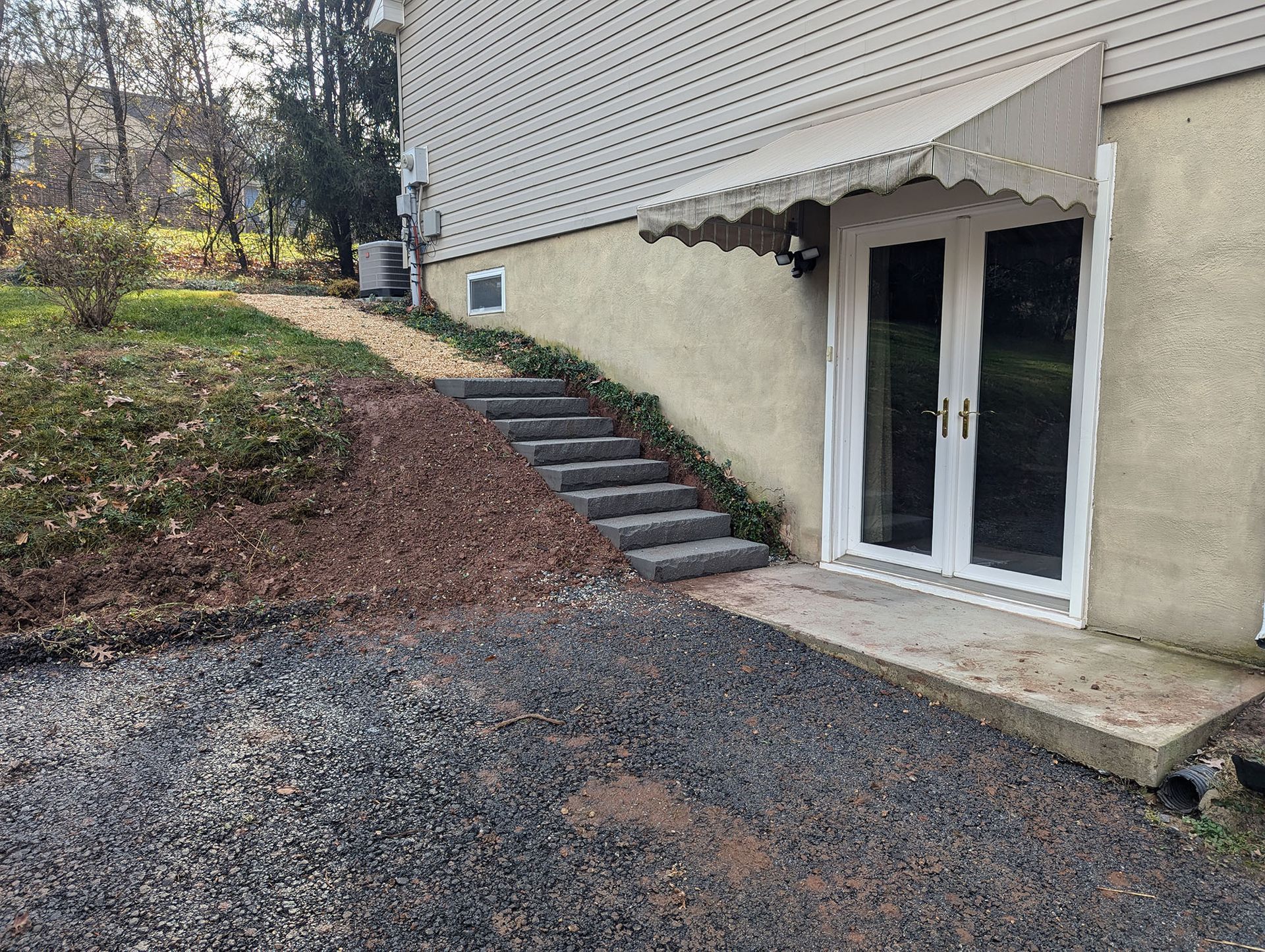 A gray gravel driveway and concrete stairs leading to a building with double doors and a tan awning.
