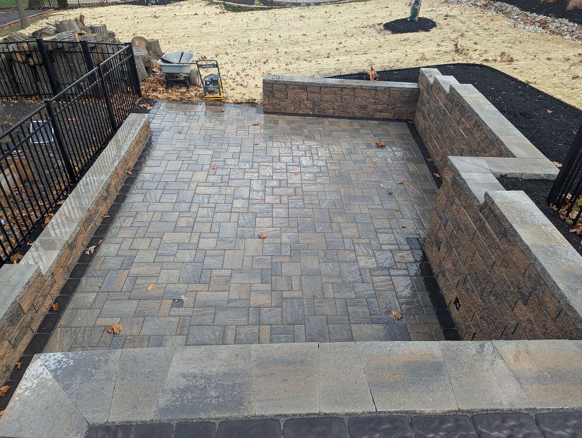 Stone patio with retaining walls, steps, and black fence. Lawn in background.