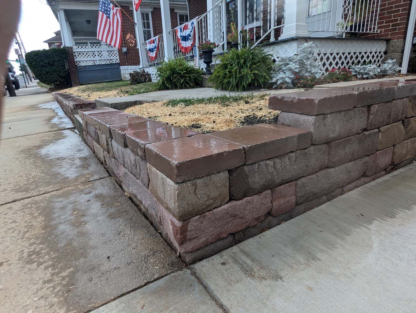Brick retaining wall in front yard with plants, wet sidewalk, and house in the background.