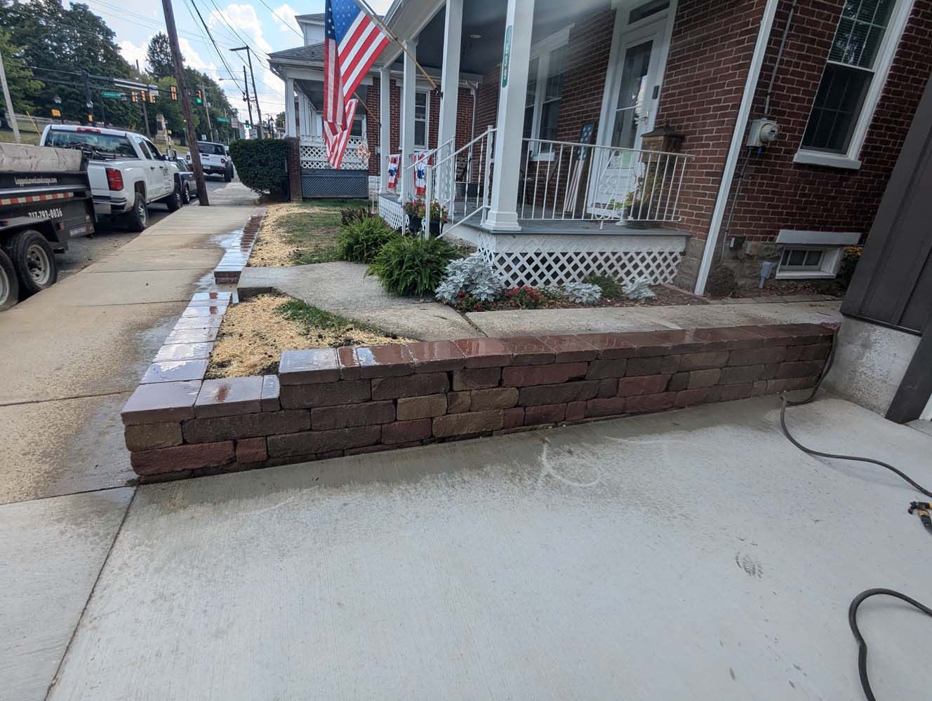 Red brick retaining wall next to a sidewalk and a brick building with a porch and American flag.