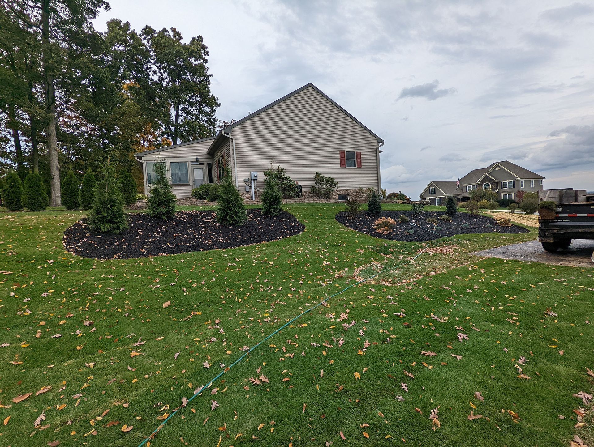 House with landscaped yard; dark mulch beds with small evergreens, green lawn, cloudy sky.