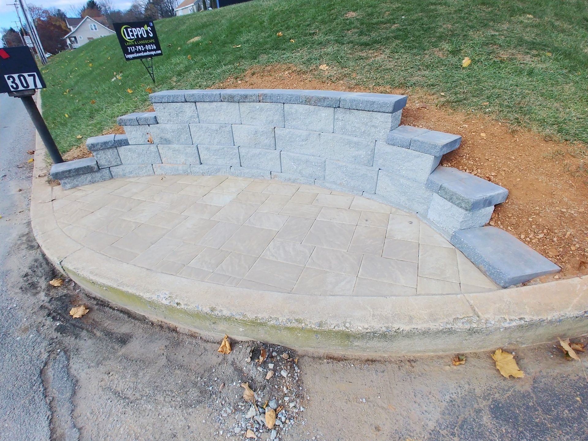 Curved stone patio and steps in a yard next to a street. Gray retaining wall, green grass, and address sign.