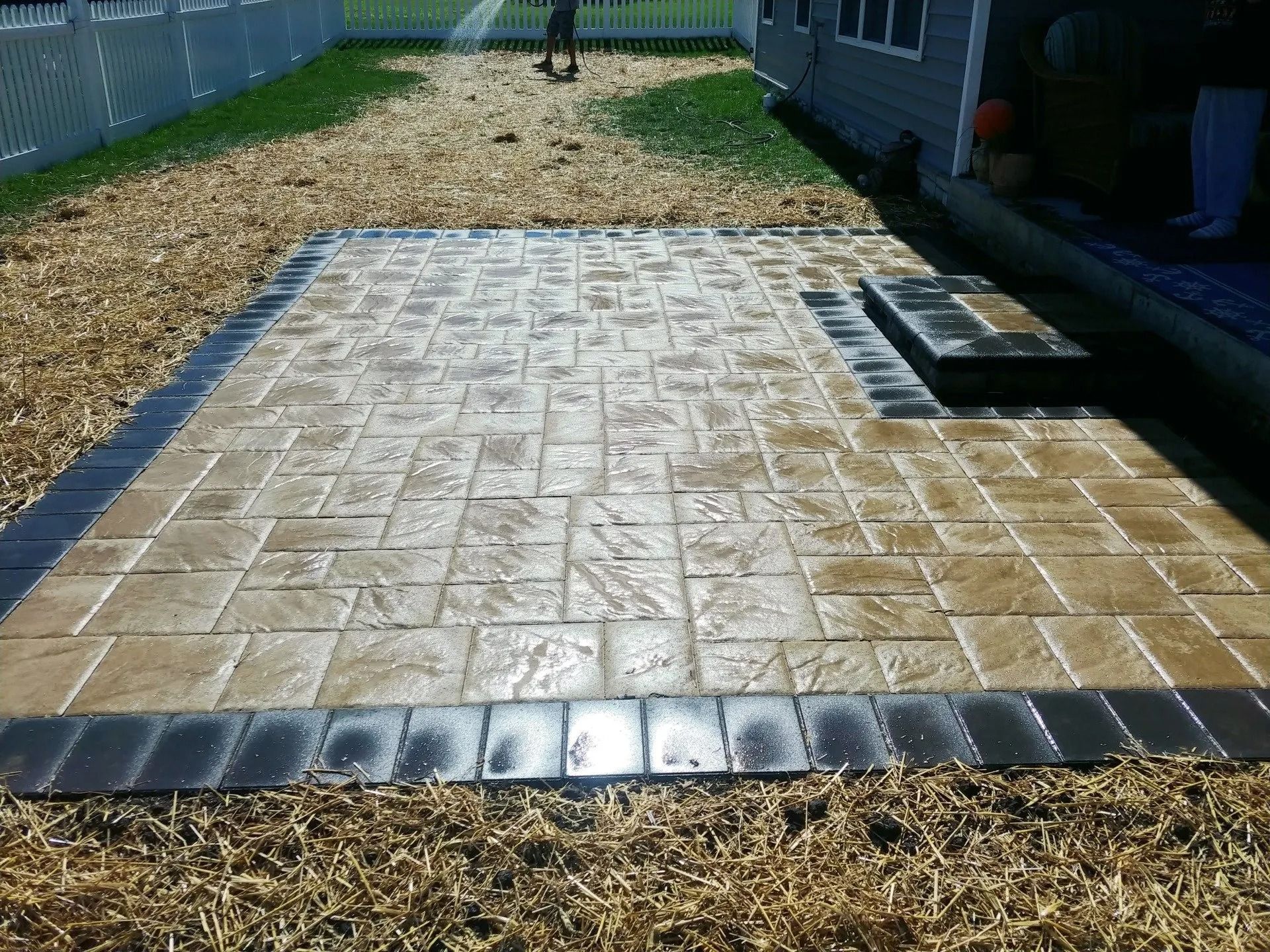 Patio with stamped concrete, surrounded by a border and mulch. A building and white fence are in the background.