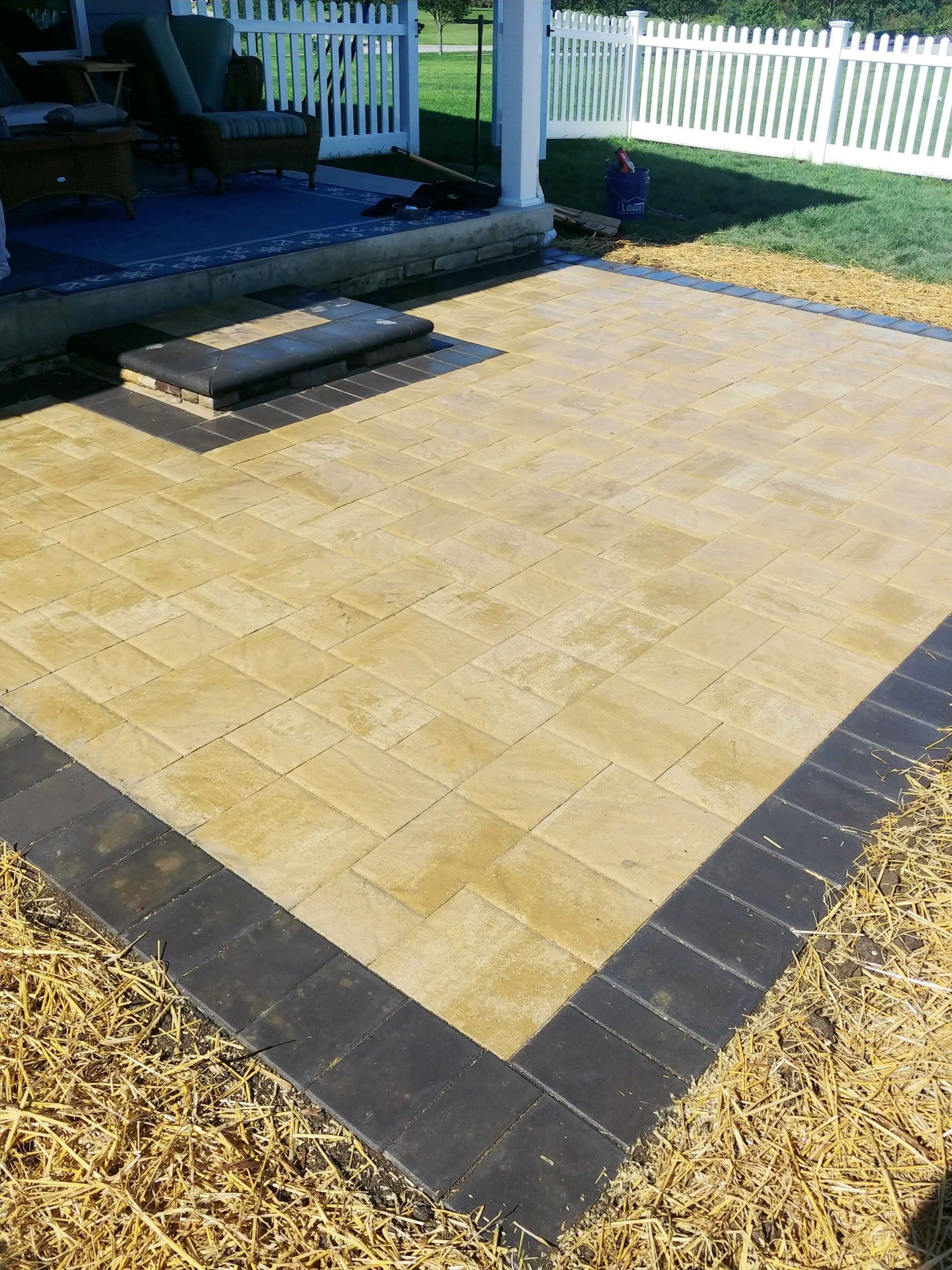 Patio with beige pavers and dark border, fire pit, white fence and grass.