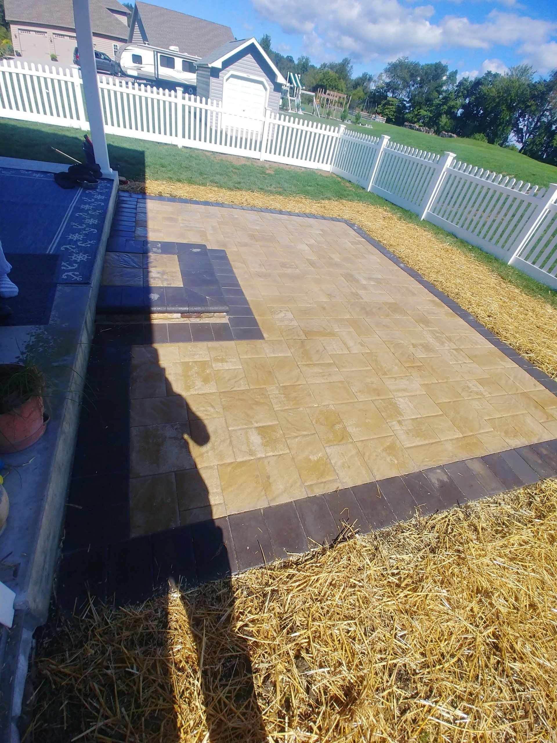 Patio with tan pavers, brown border, next to a white picket fence and lawn with tan gravel.