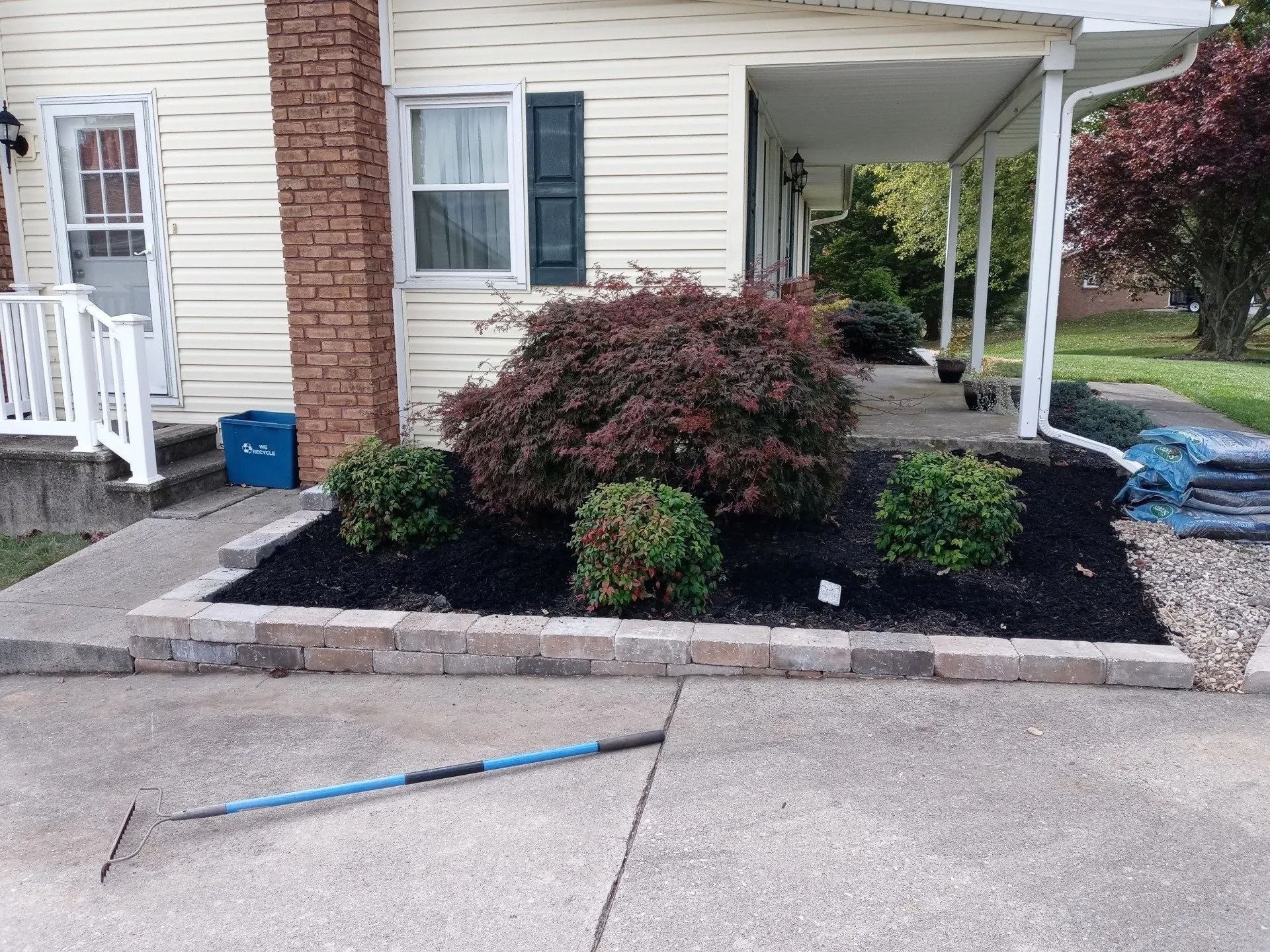 A house with a small garden bed outlined with blocks, featuring a large red-leaved bush and several green shrubs.