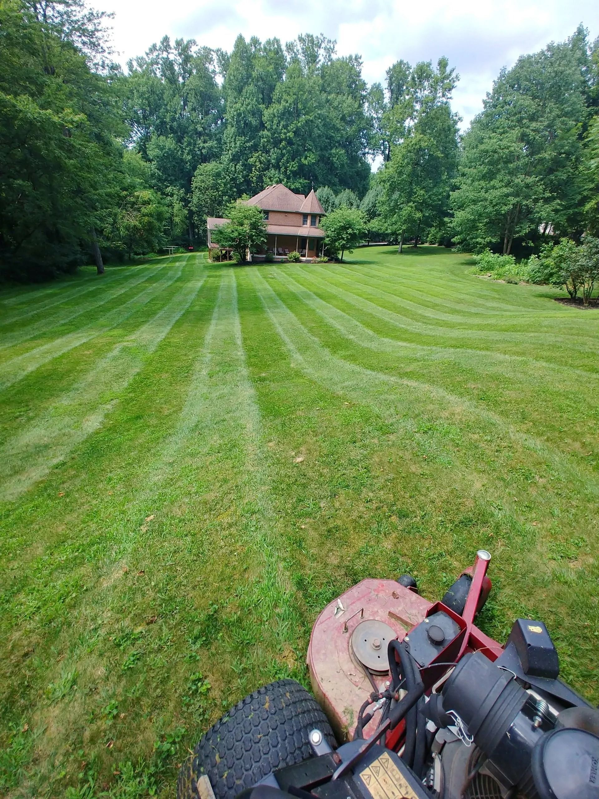 Lawn mower cutting stripes in a large green yard, a house in the background on a sunny day.