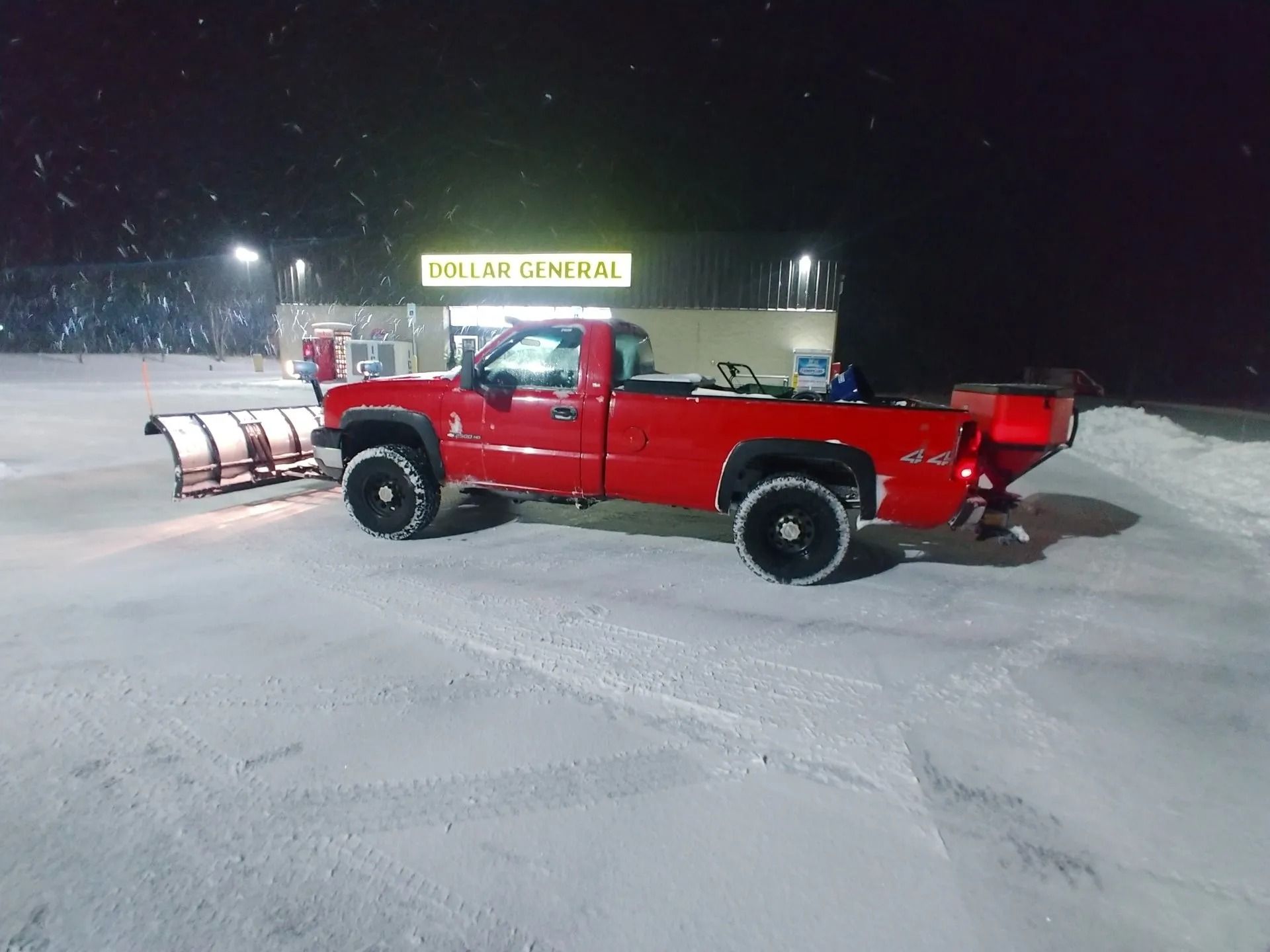 Red pickup truck with snowplow and salt spreader plowing snowy parking lot in front of Dollar General store at night.