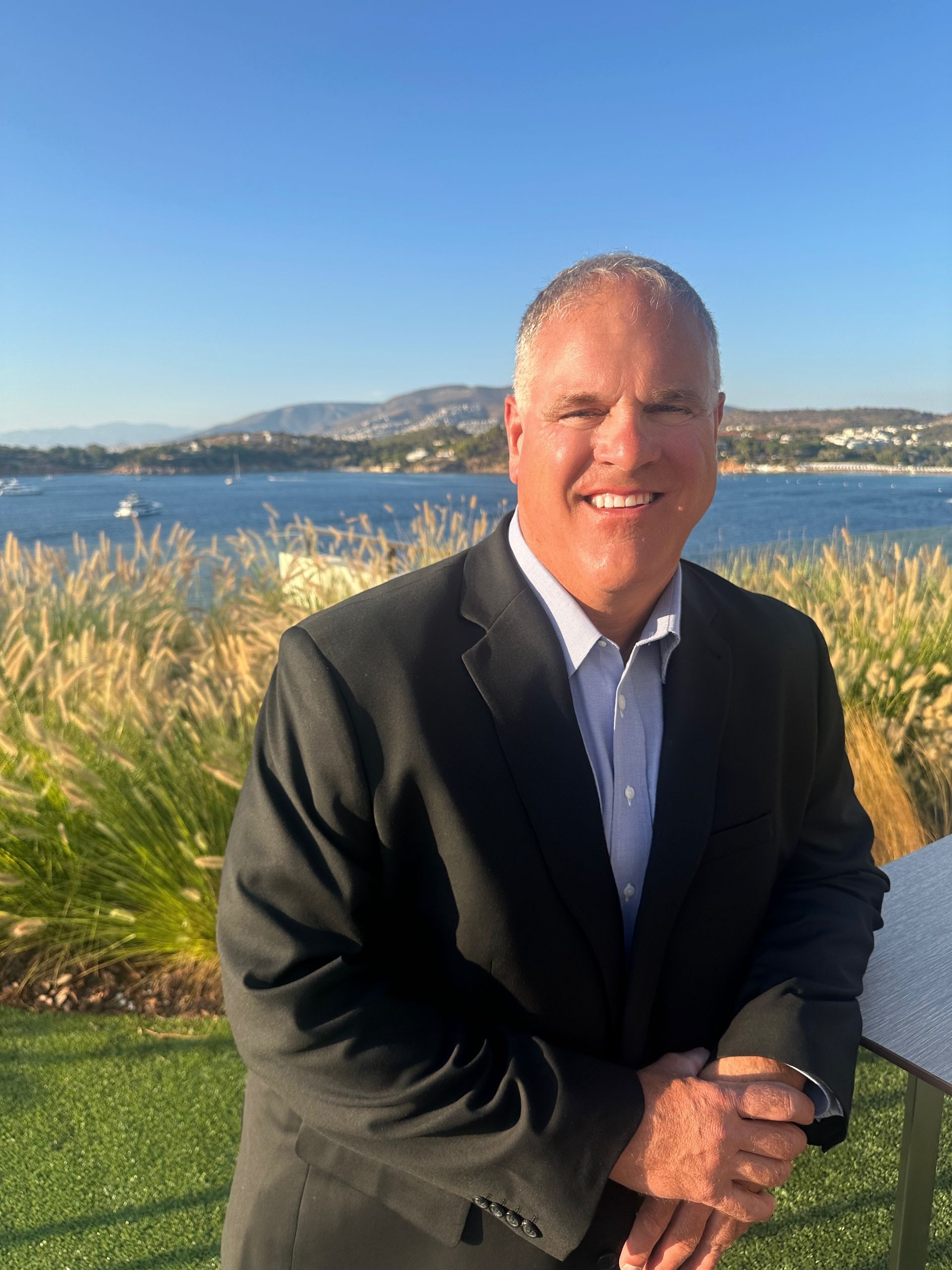 Smiling man in a black blazer stands by a waterfront with grass, hills, and blue sky in the background