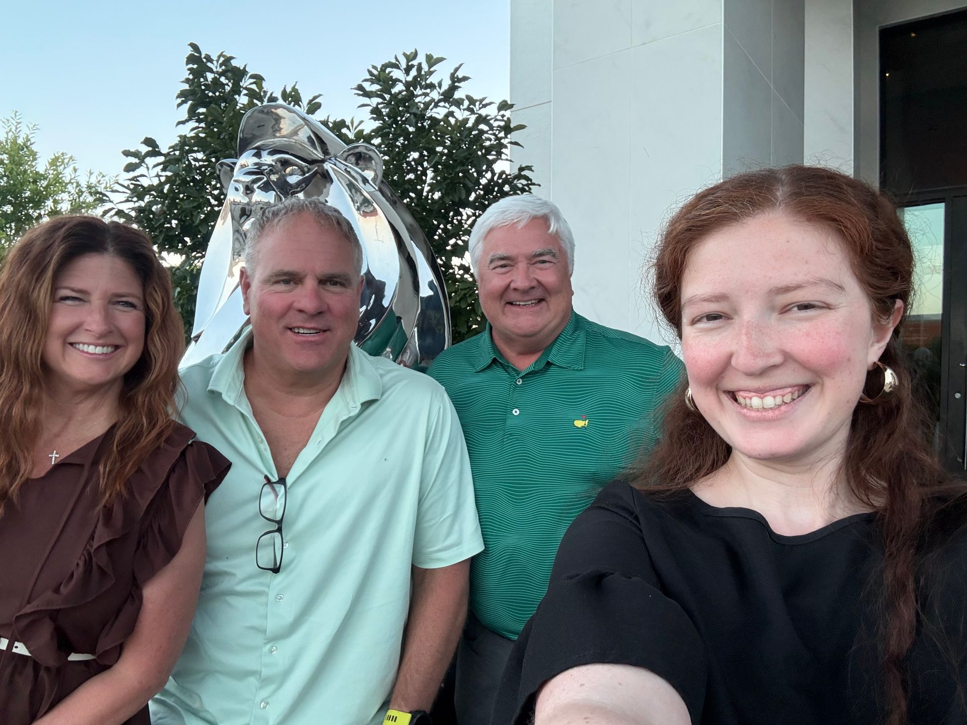 Four people smiling outdoors, standing together near a building entrance.
