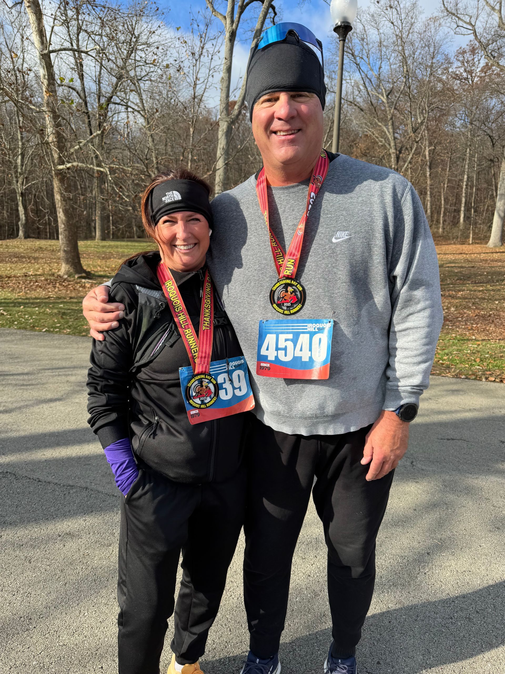 Two runners smiling outdoors, wearing race bibs and medals, with arms around each other.