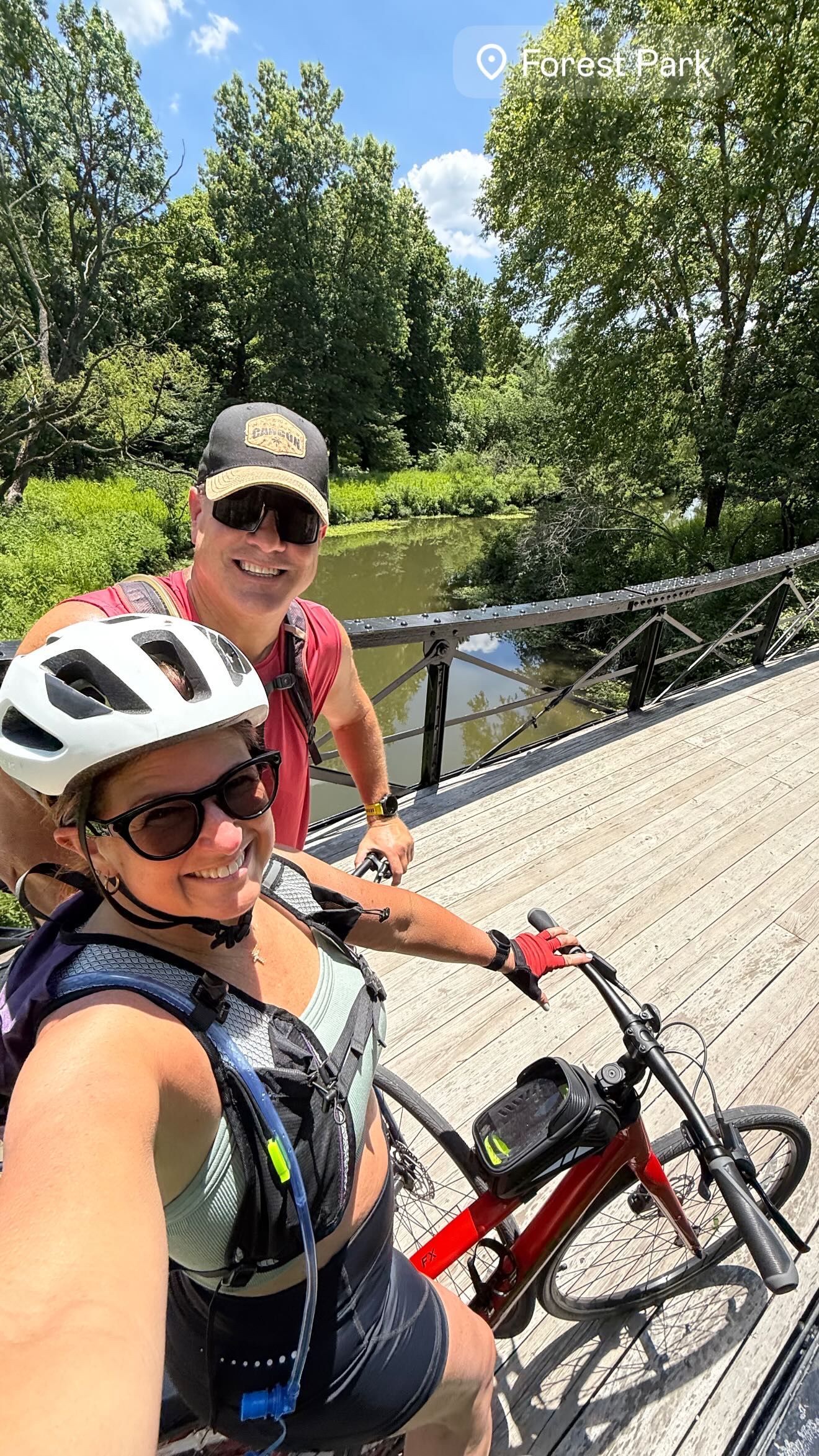 Two cyclists taking a selfie on a bridge beside a green river under sunny trees