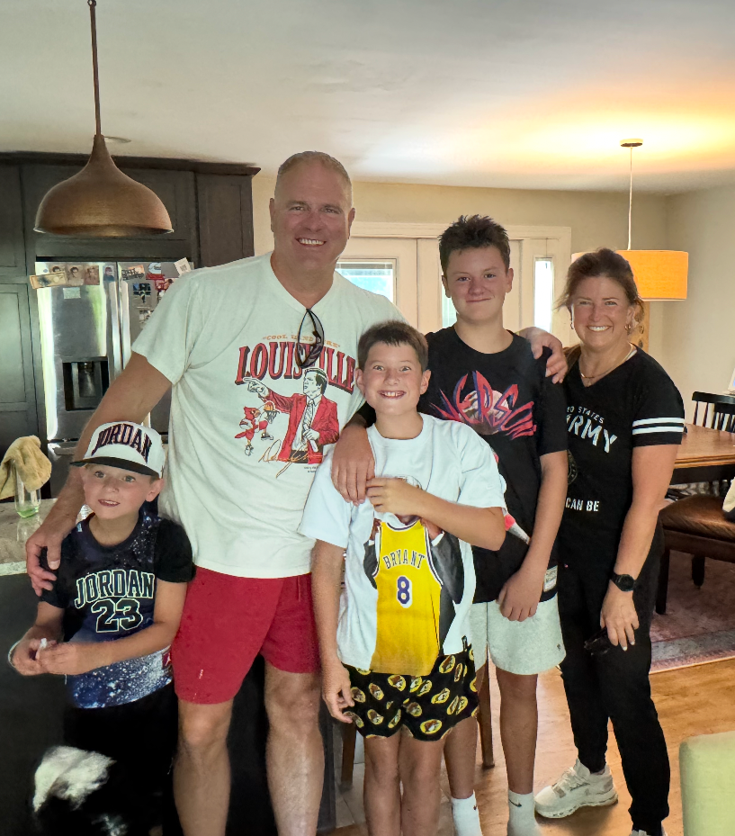 Family of five smiling indoors in a kitchen and dining area, standing close together.
