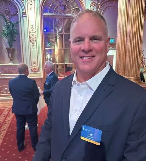 Smiling man in a navy suit with a name badge standing in an ornate hall with red carpet and gold columns