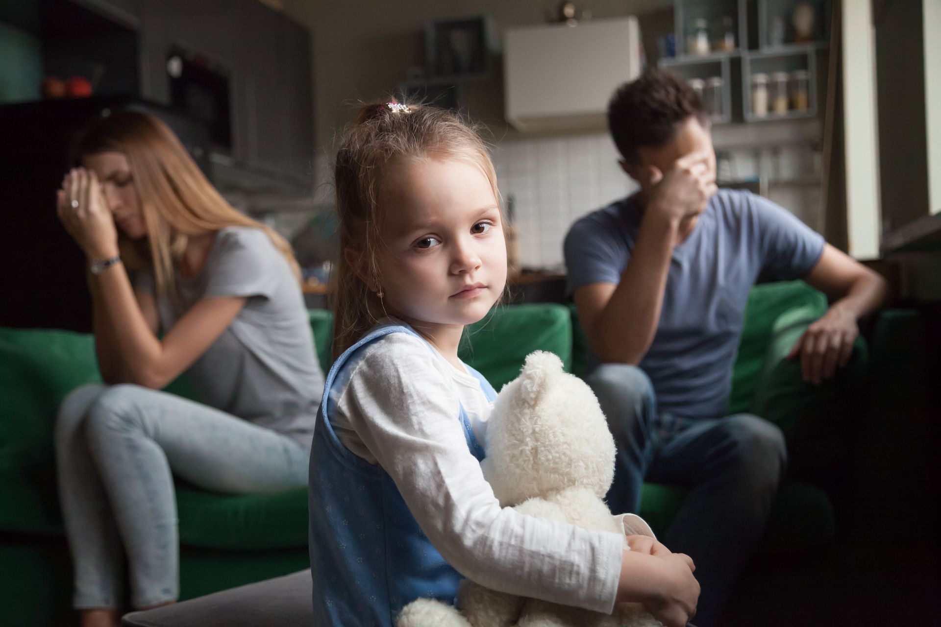 Sad child holding teddy bear, parents sit separately, covering faces.